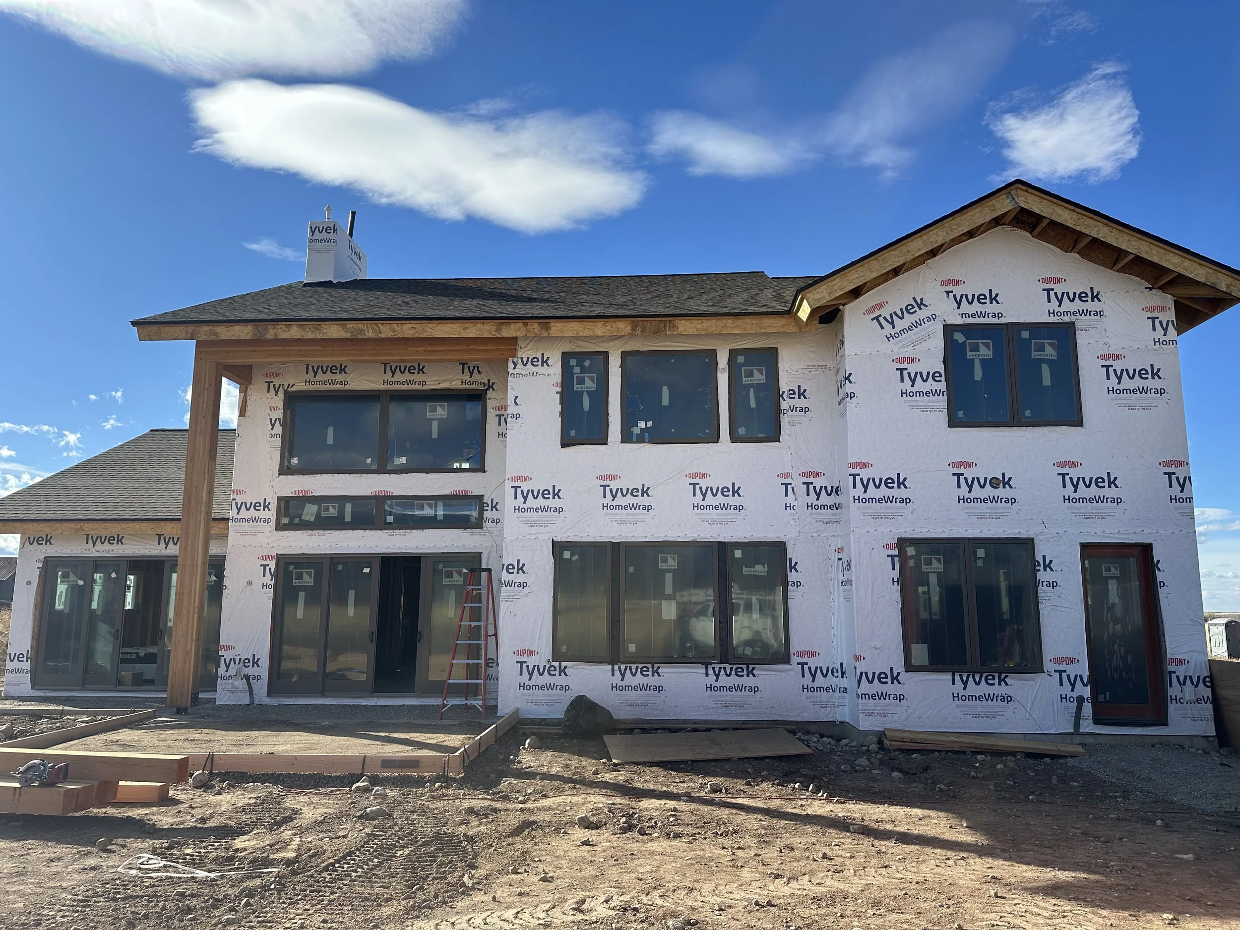 Two-story house under construction with Tyvek HomeWrap and large windows, dirt ground in front, and a blue sky with clouds overhead.