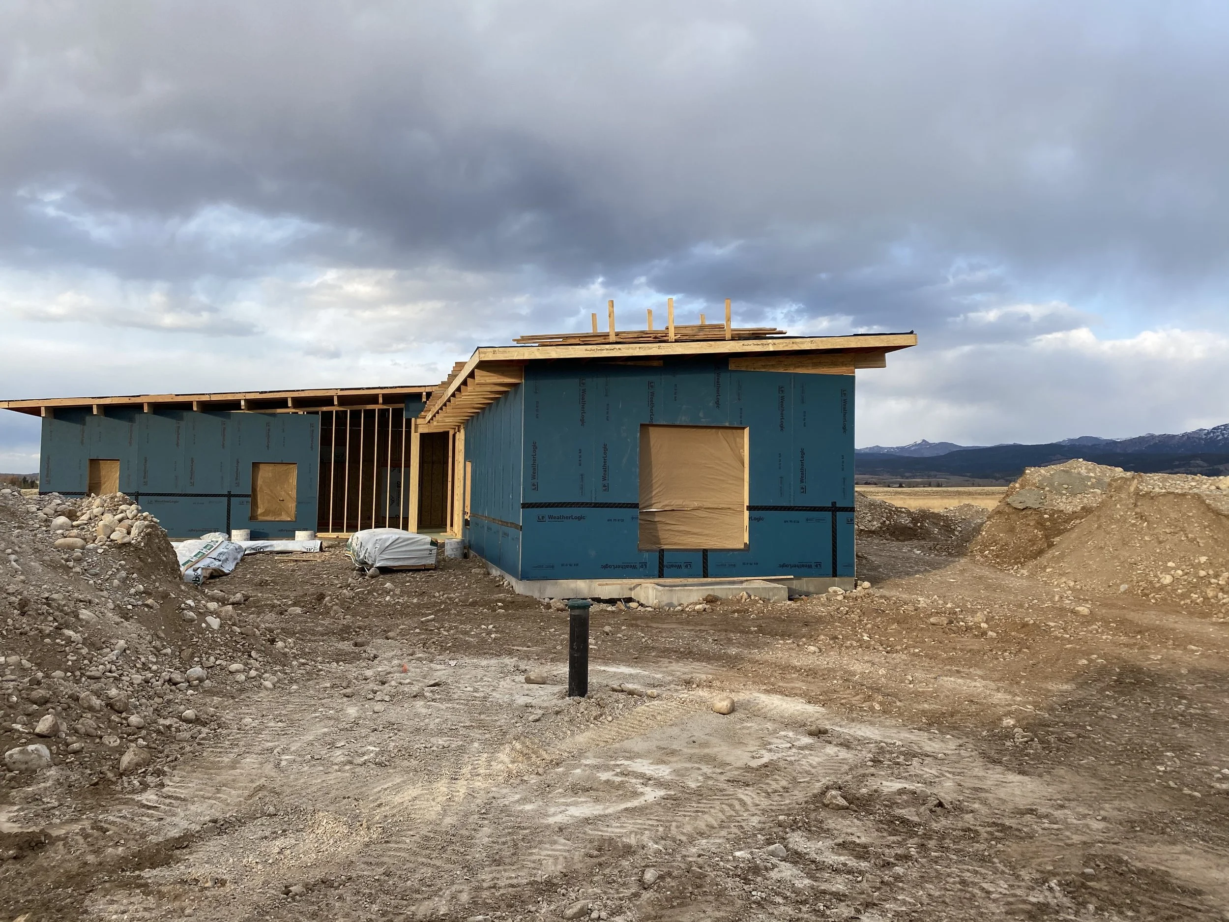 Under construction house with blue sheathing in a rural area, dirt ground, and mountainous landscape in the background.