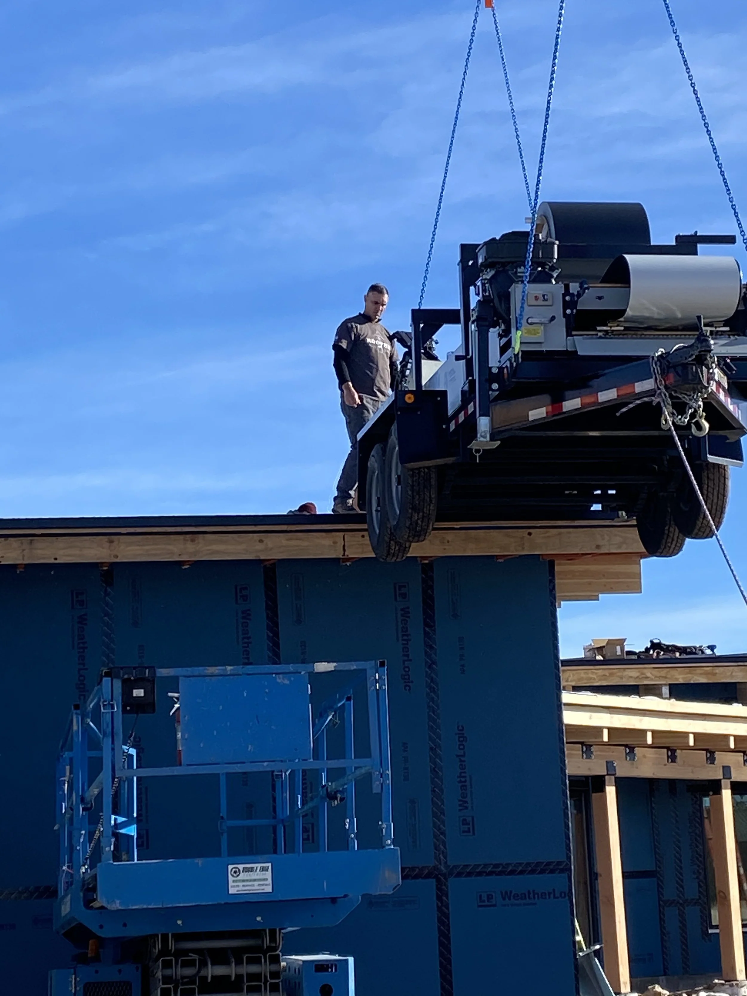 A construction worker standing on a wooden roof under construction, next to a suspended platform, with a blue sky in the background.