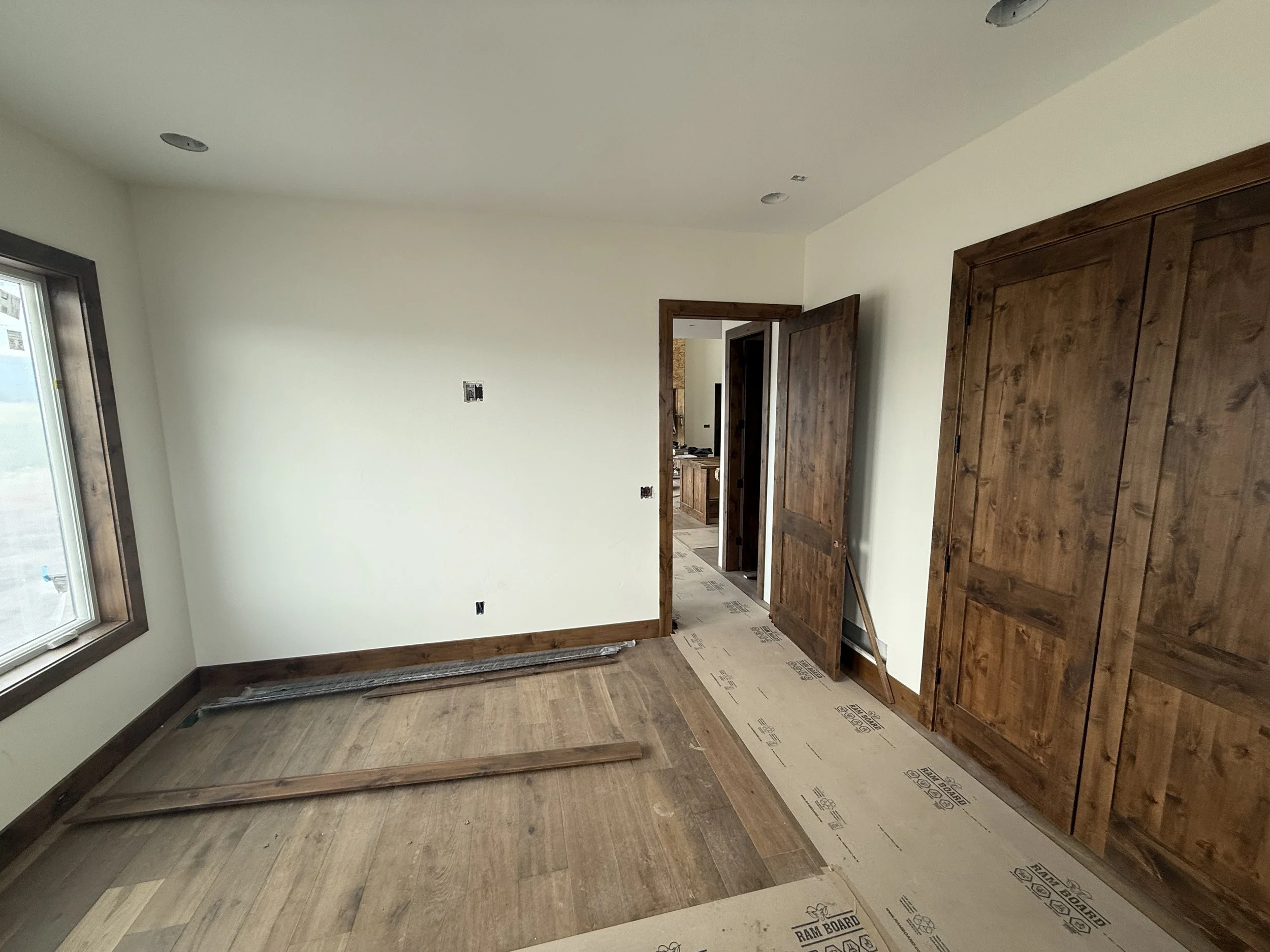 Empty room with unfinished flooring, white walls, wooden trim, and a large window. The room appears to be under construction or renovation with some wood planks on the floor and uninstalled hardware near the doorway.