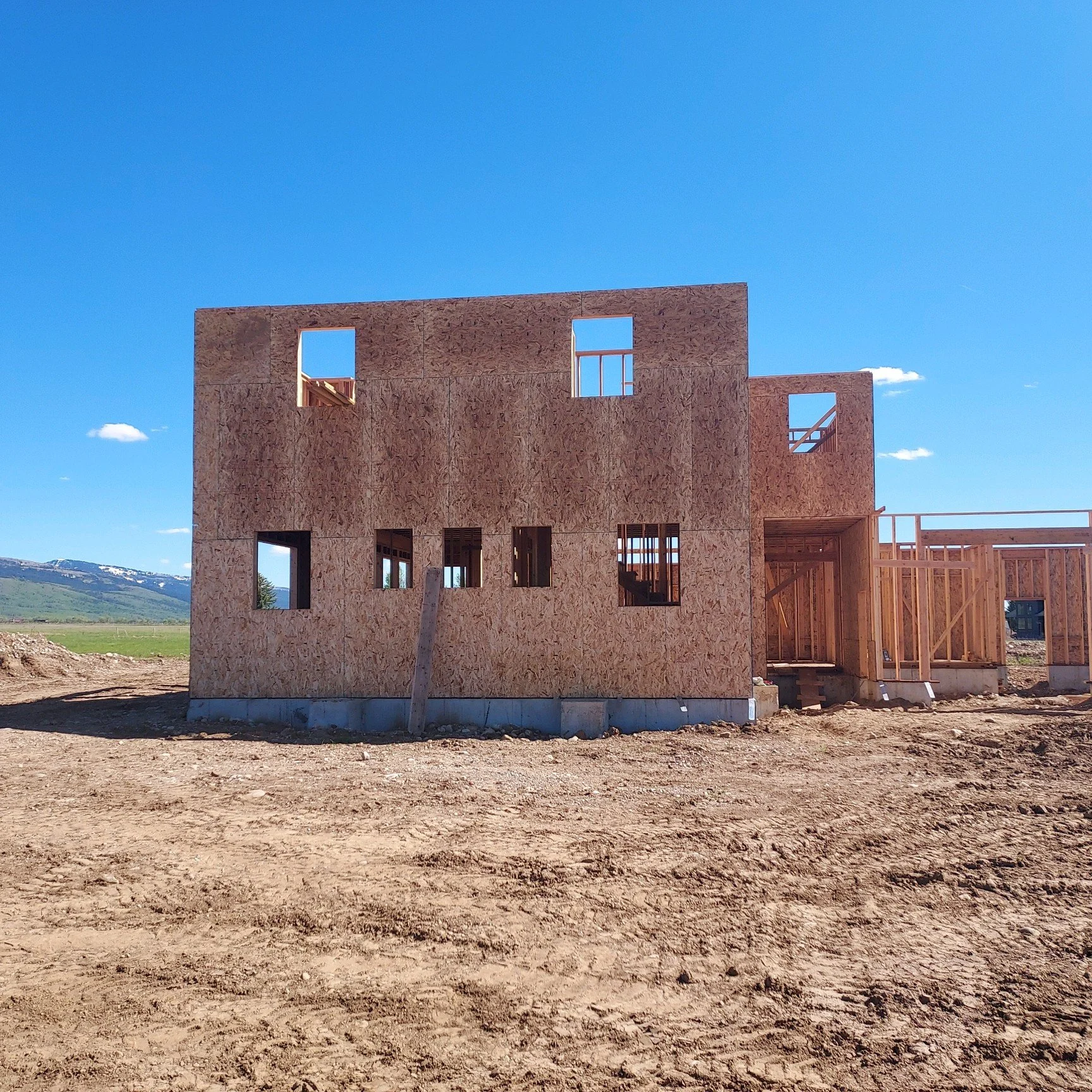 Under construction wooden house frame on a dirt lot with clear blue sky.