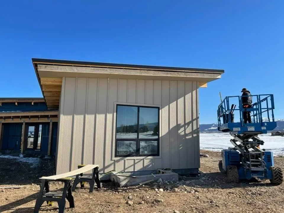 Construction worker on a blue lift working on the exterior of a beige house with vertical siding, a large window, and a sloped roof in a cold, snowy landscape.