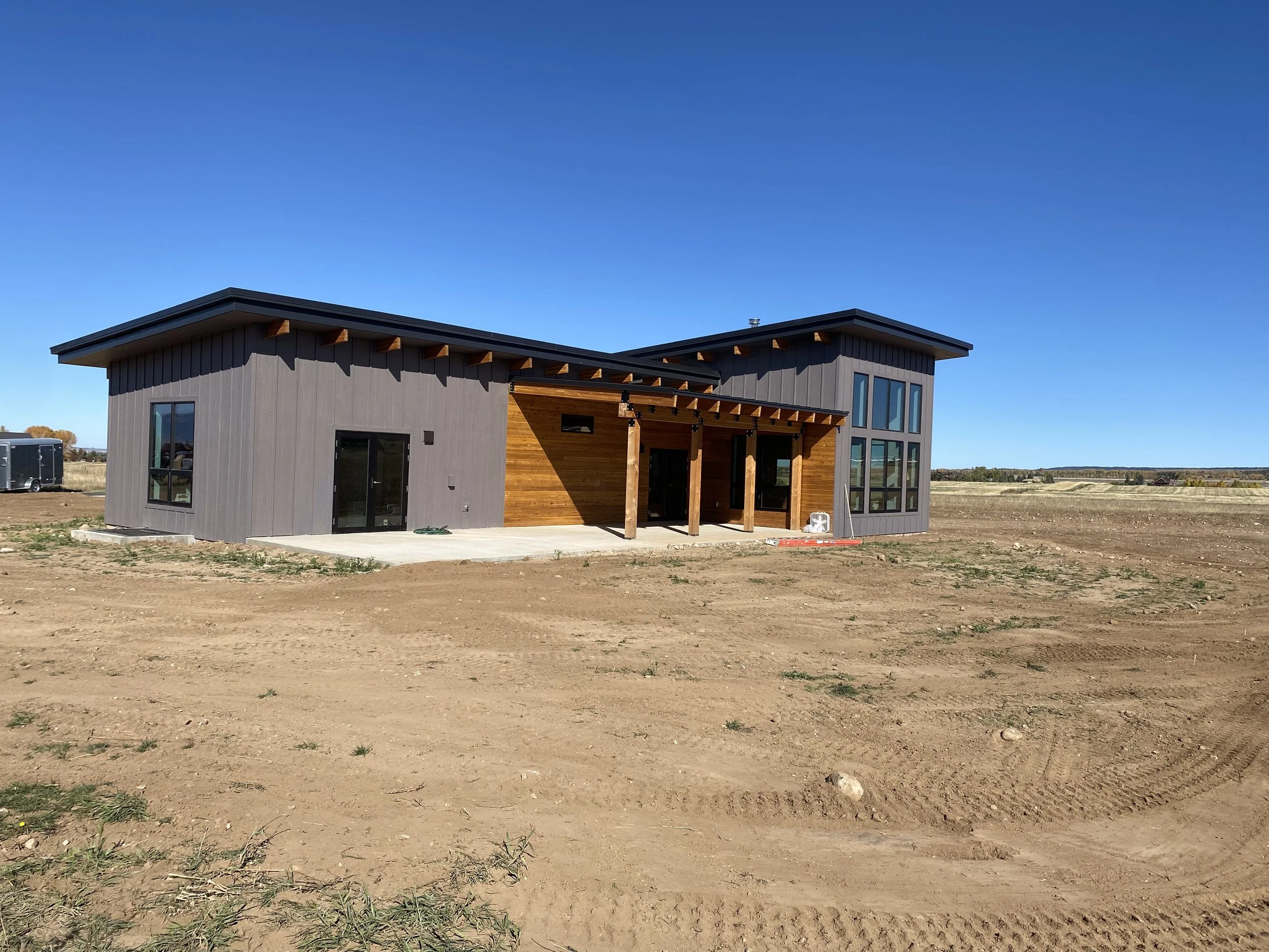Newly constructed modern house with gray siding and wooden accents, set on a flat, bare lot under clear blue sky.