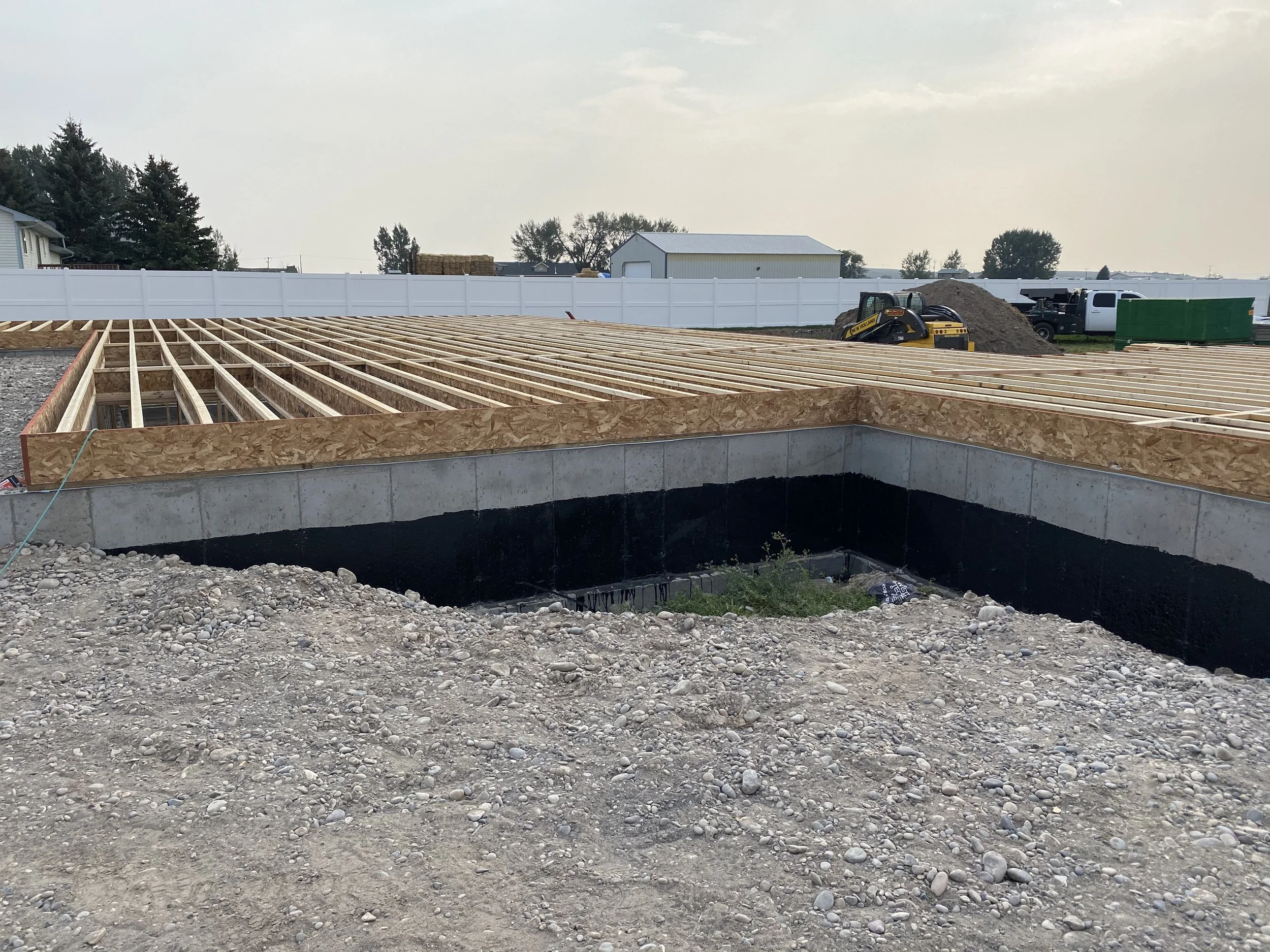 Construction site with foundation and wooden framing for a building, gravel in foreground, a white fence in the background, and a small construction vehicle near a pile of dirt.