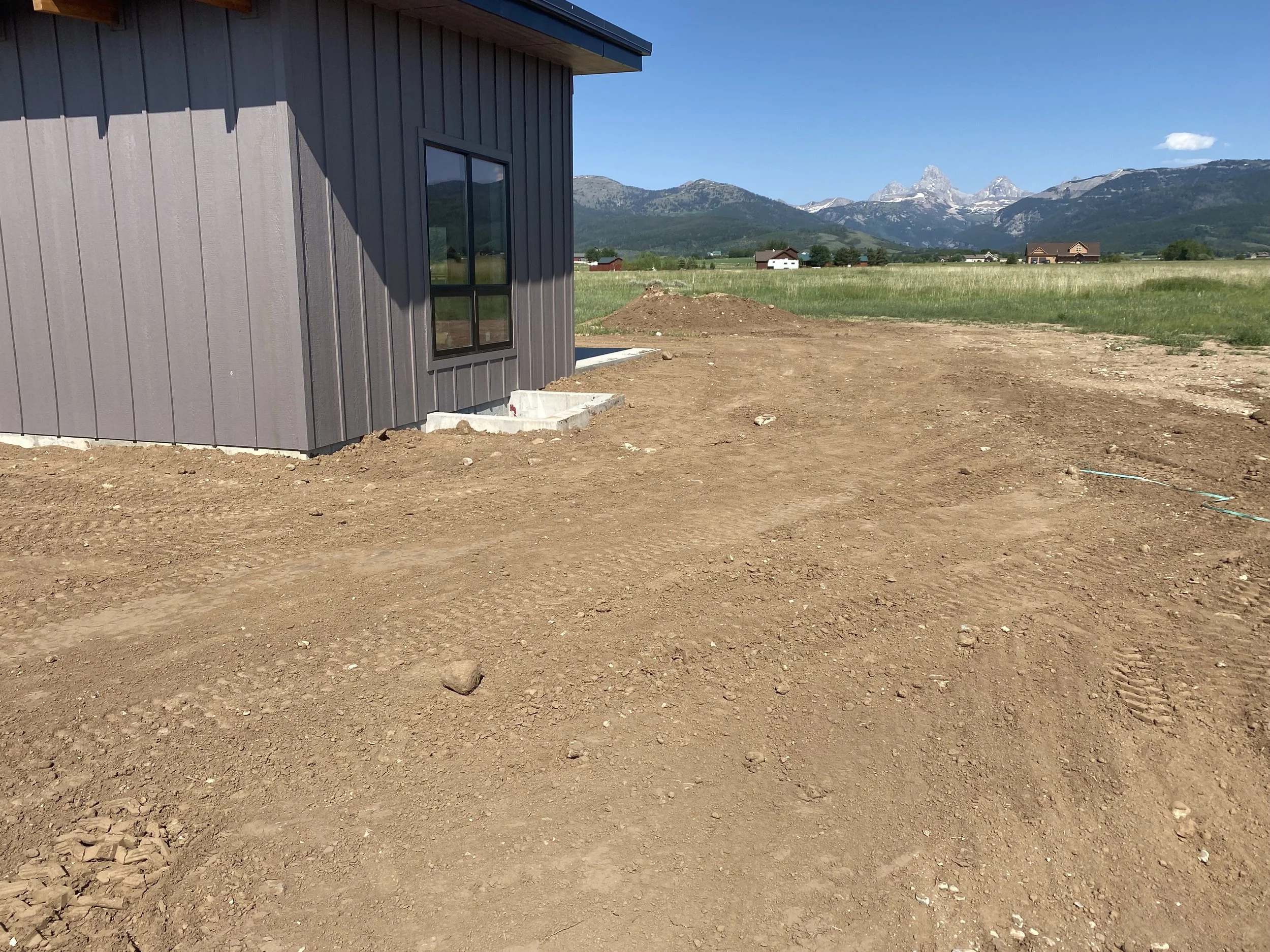 Modern custom home exterior with clean lines and mountain landscape in Eastern Idaho.
A dirt yard next to a gray house with a black window, with a mountain range in the background under a clear blue sky.