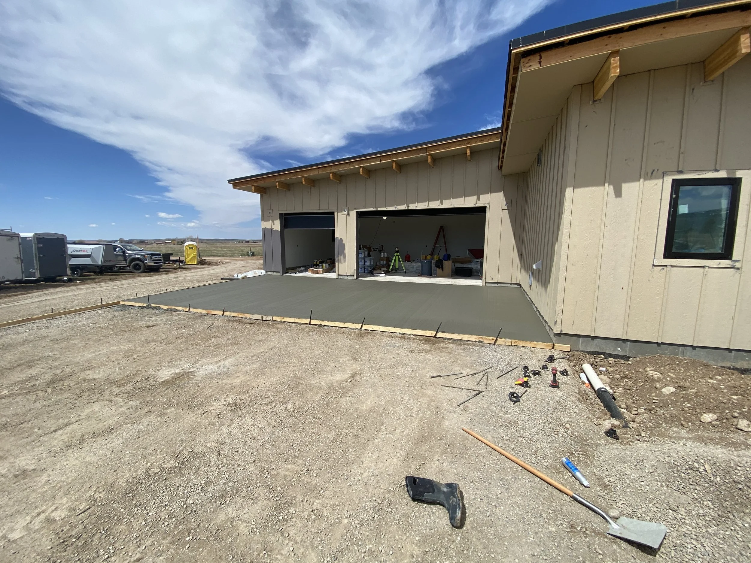 Newly poured concrete slab in front of a garage under construction, with construction tools and equipment scattered around, and a partly cloudy sky overhead.