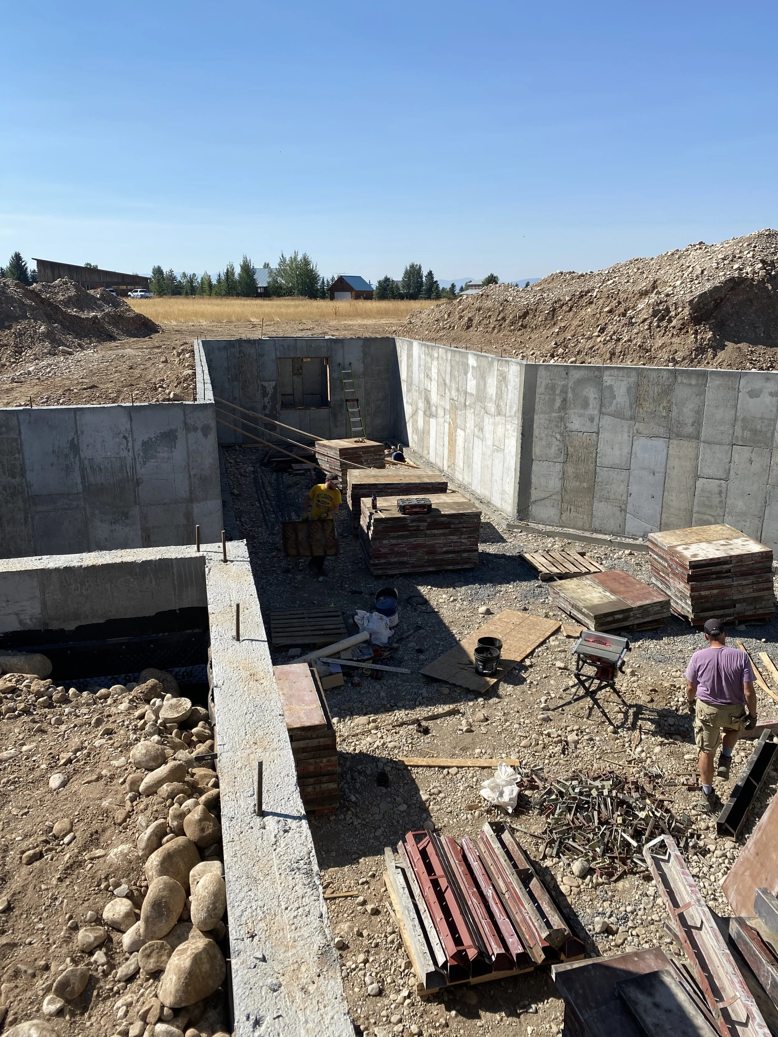 Construction site with workers building concrete walls, piles of bricks, tools, and handrails in a rural area on a sunny day.