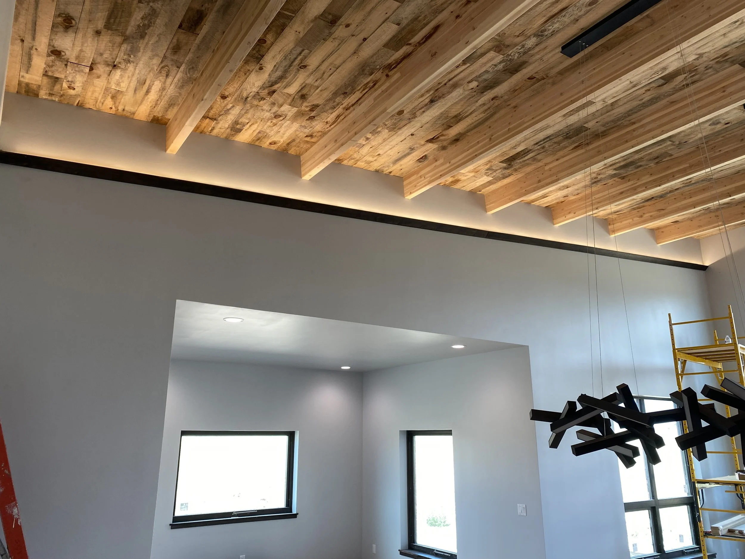 Interior of a modern house under construction with a wood-paneled ceiling, white walls, recessed lighting, large windows, and a yellow scaffold.