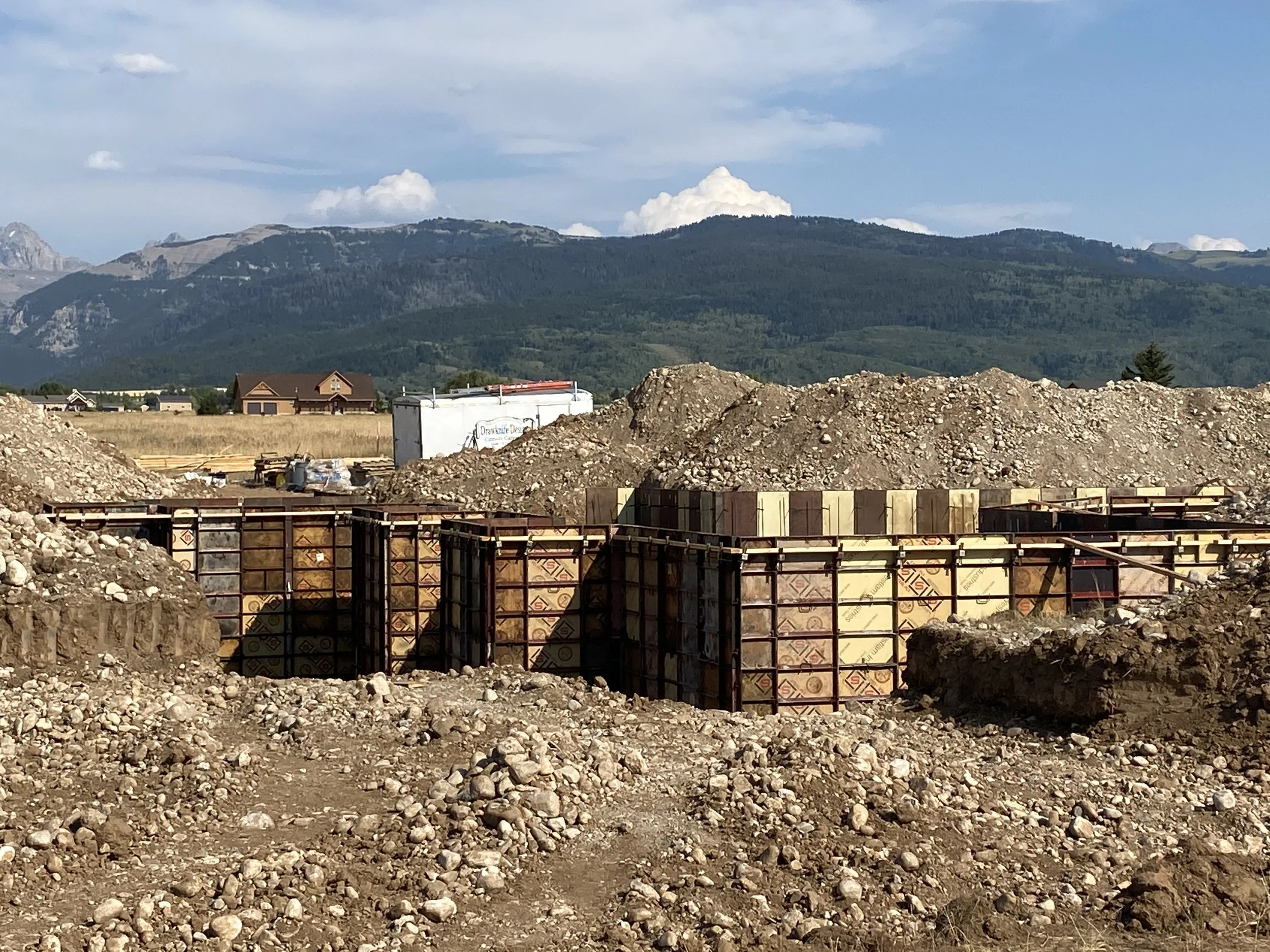 Construction site with foundation forms and dirt piles, mountains and blue sky in the background.