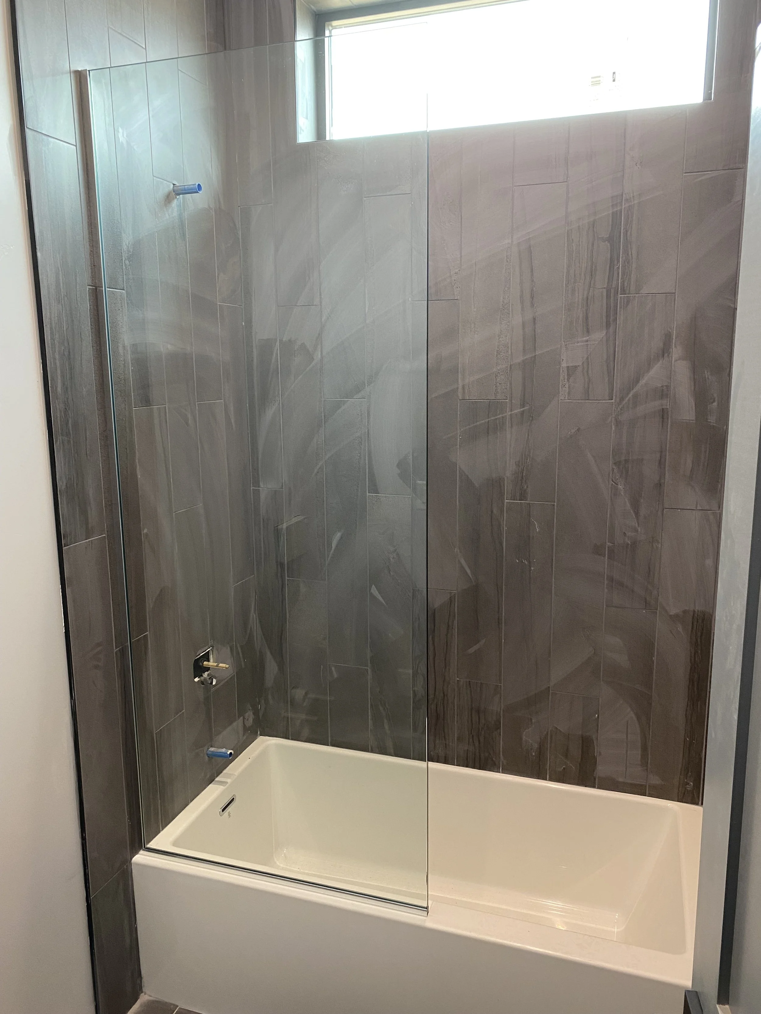 A modern shower with dark gray tiled walls, a glass shower door, and a small frosted window.