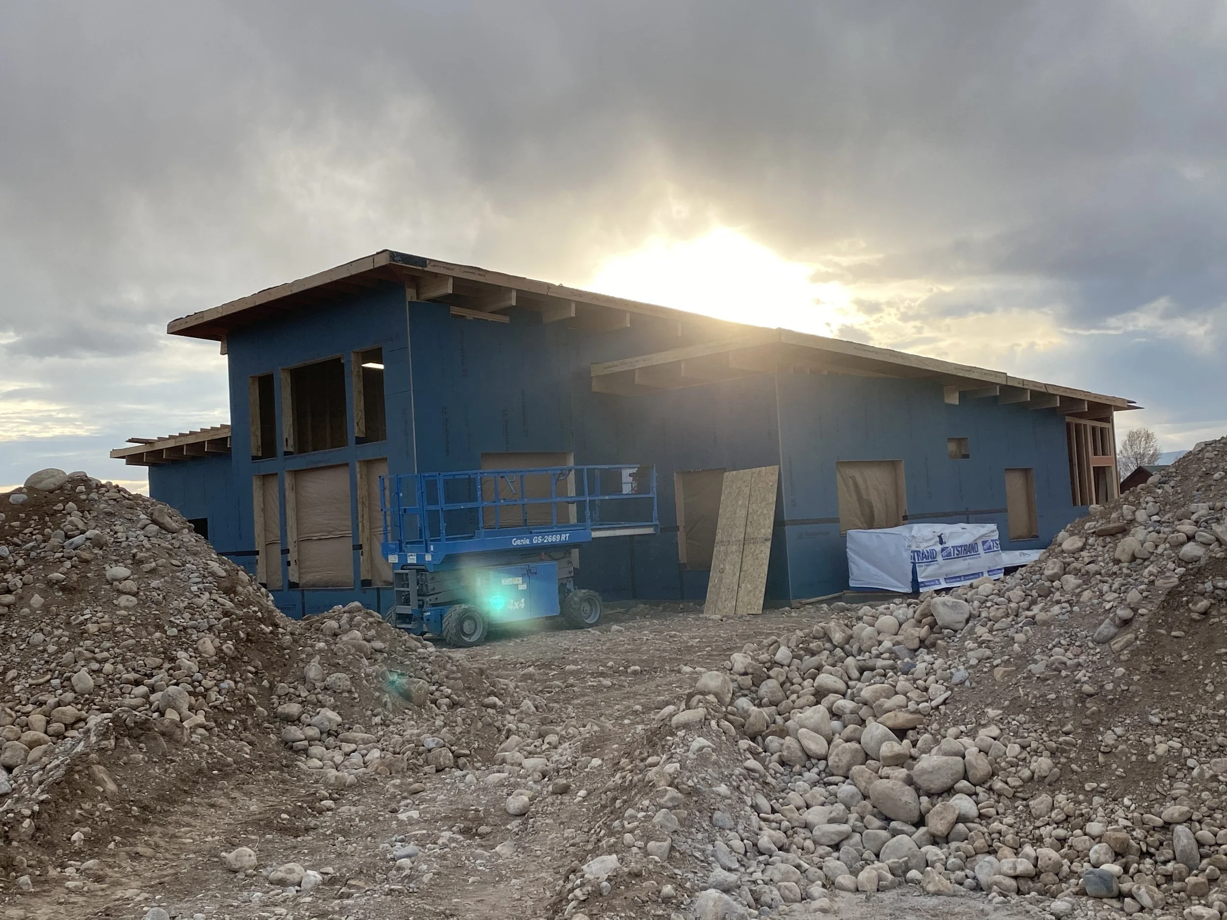 A house under construction with blue exterior sheathing, surrounded by dirt and rocks, with a construction lift nearby, against a backdrop of a cloudy sky and a setting or rising sun.