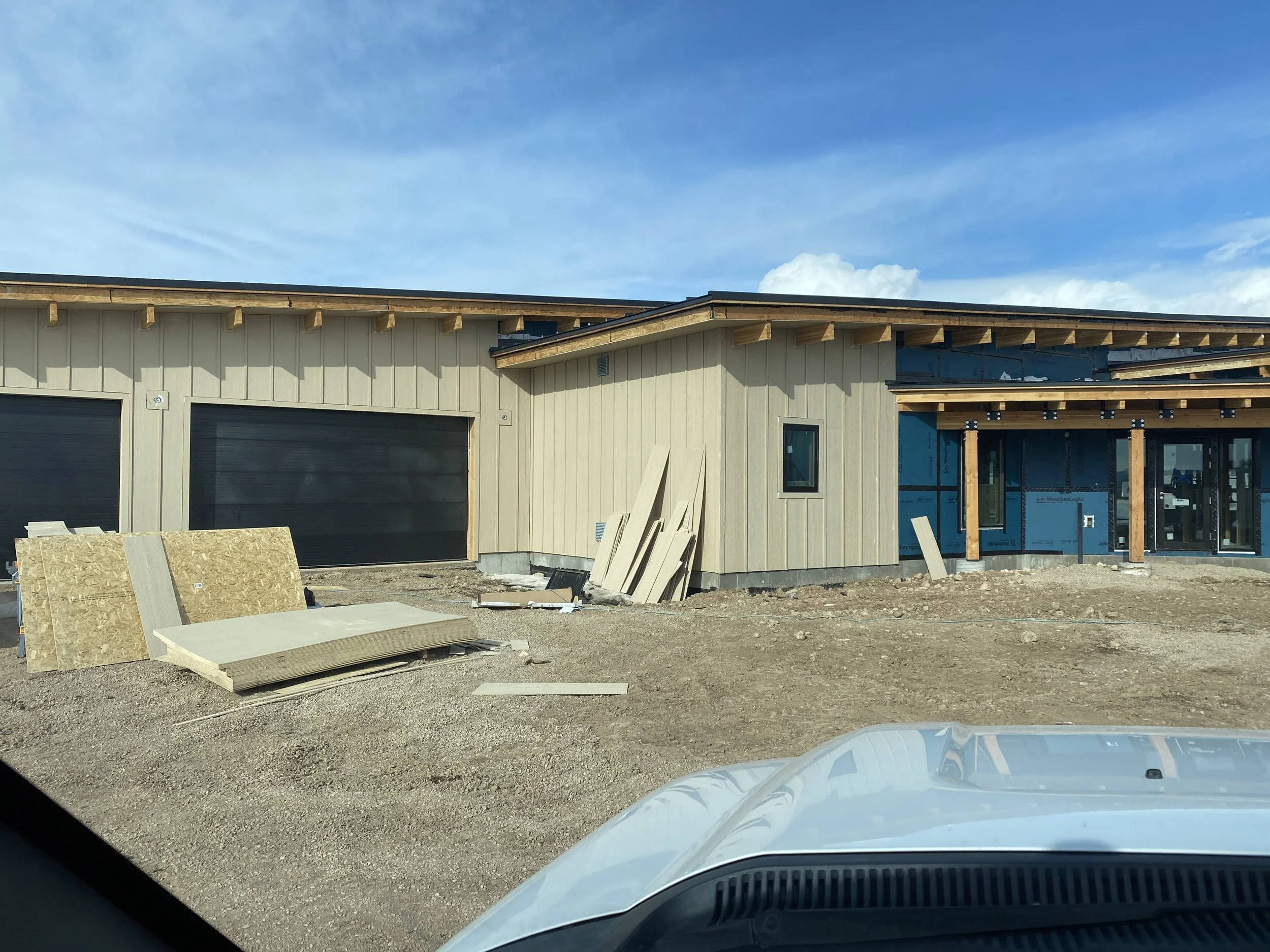 Construction site of a house with beige siding, black garage doors, and exposed wooden beams and columns, on a dirt lot with building materials in the foreground under a blue sky.