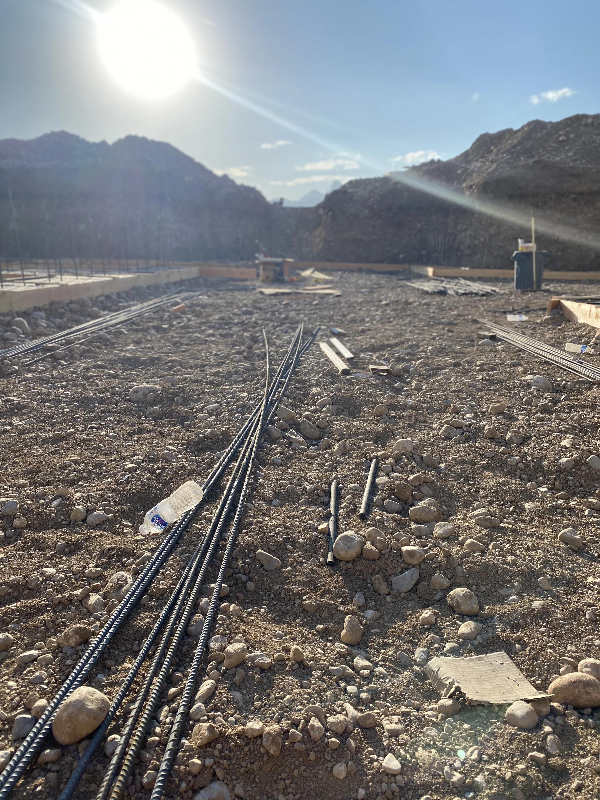 Construction site with steel rebar, rocks, and construction materials scattered on dirt ground, with mountains and clear sunny sky in the background.