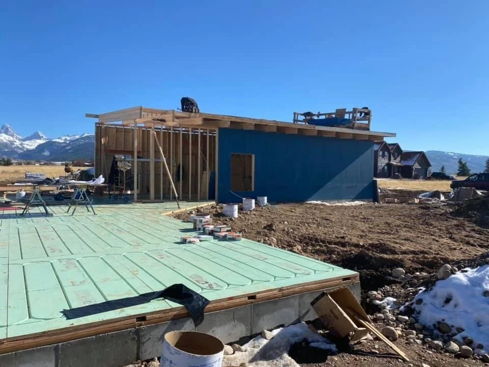 Construction site of a house with wooden framing and partially completed blue exterior wall, set in a snowy, mountainous landscape on a clear day.
