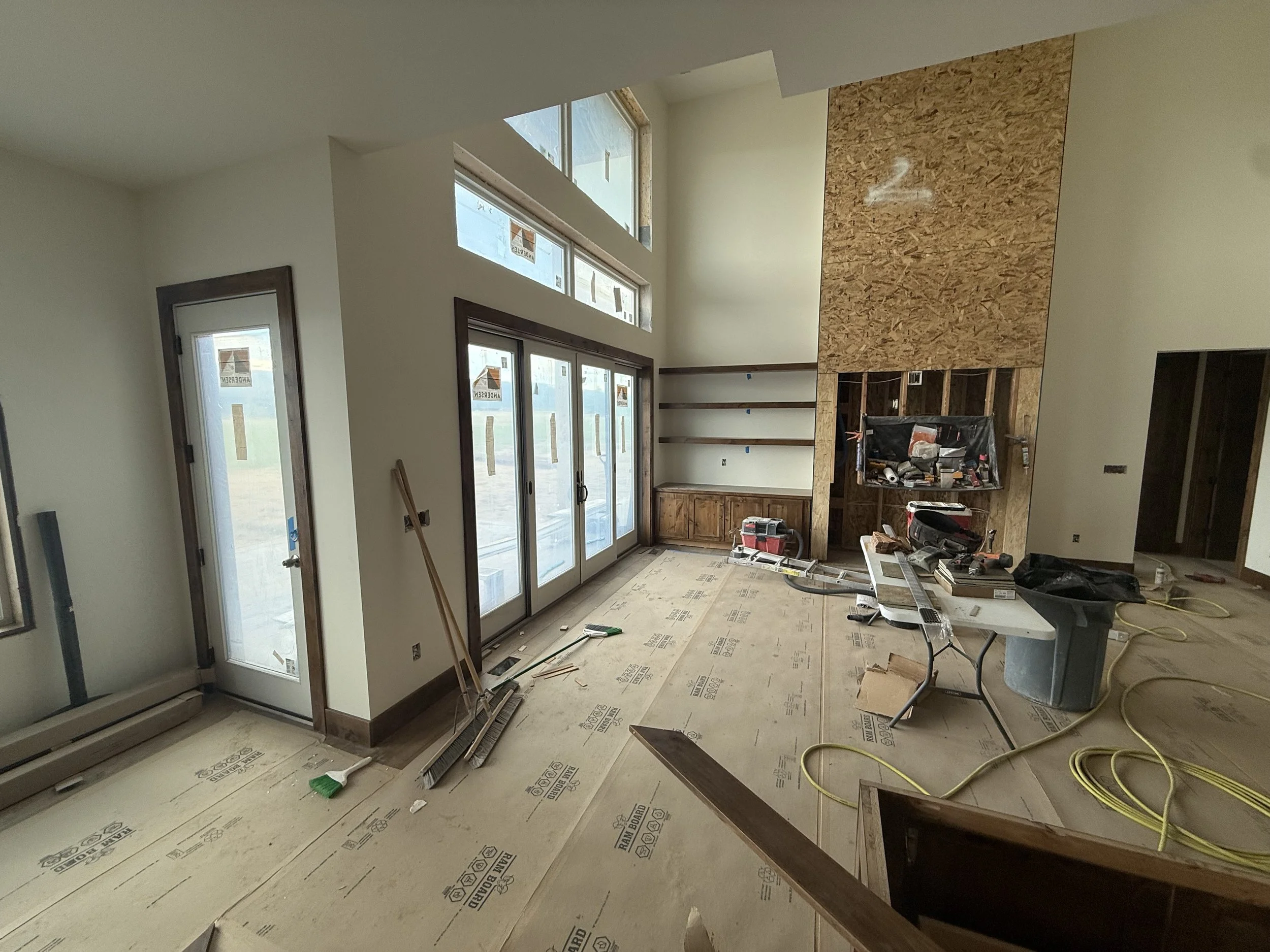 A room under construction with large windows and doors, unfinished walls, with construction tools and materials scattered around, including a sawhorse, power tools, and yellow extension cords.