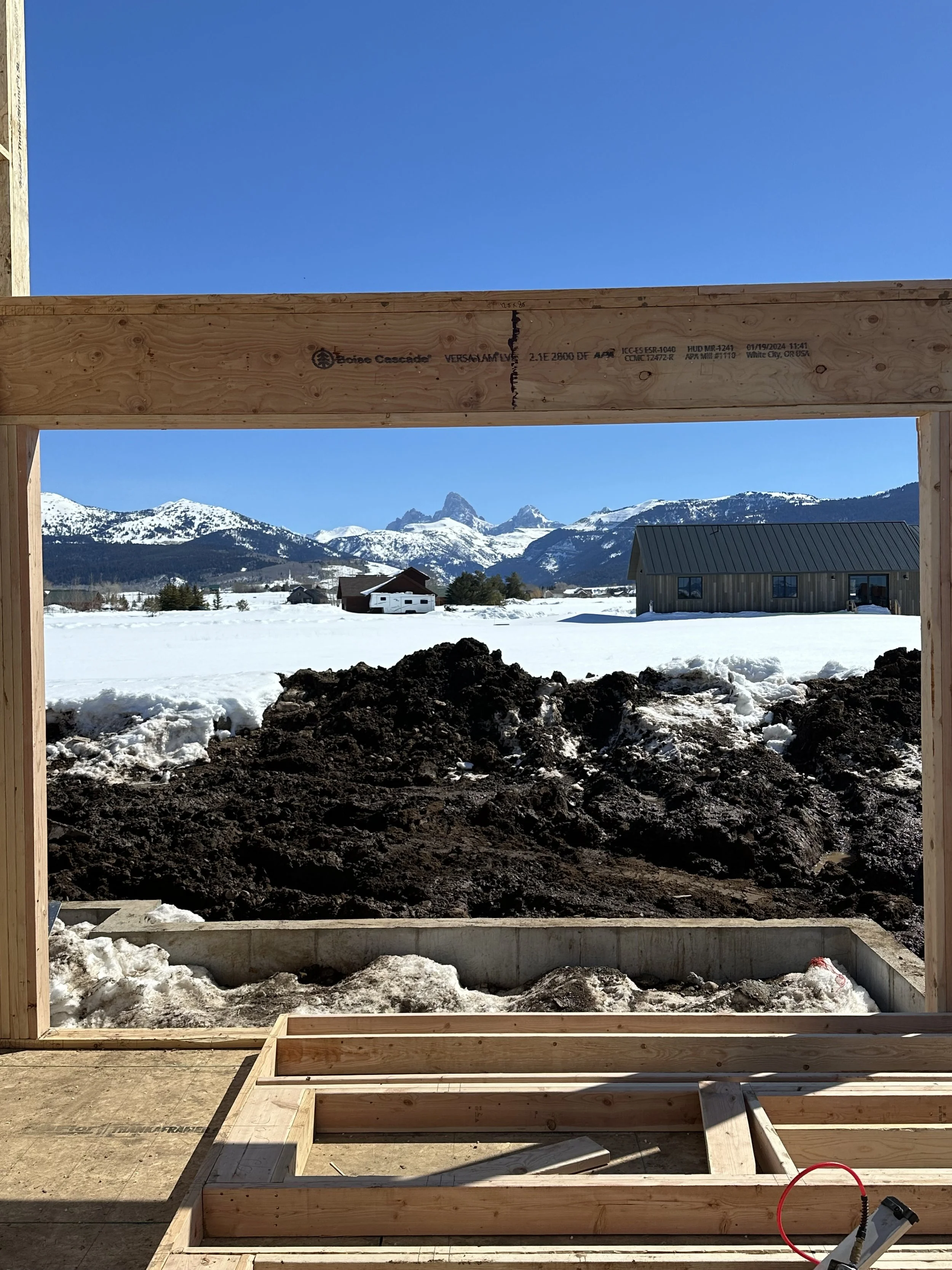 View from a building under construction with wooden framing, looking out across snow-covered ground and mountains in the distance under a clear blue sky.