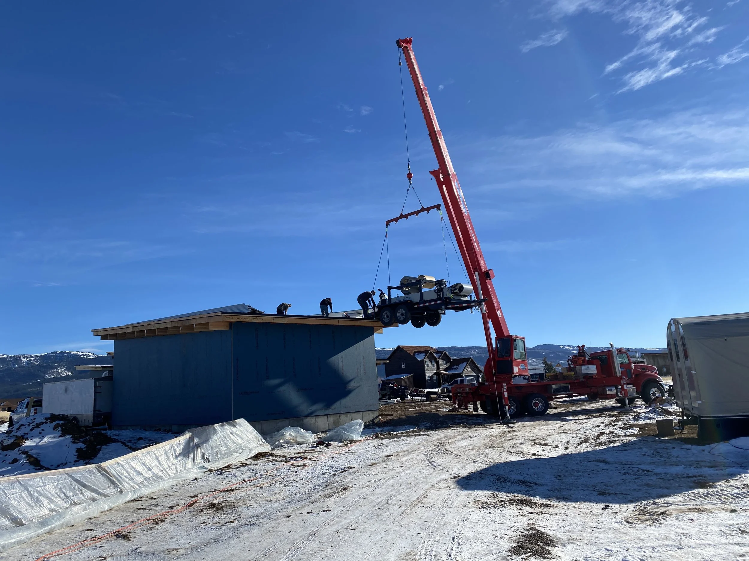 A construction site with a large red crane lifting construction equipment onto the roof of a building under construction, with snow-covered ground and mountains in the background.