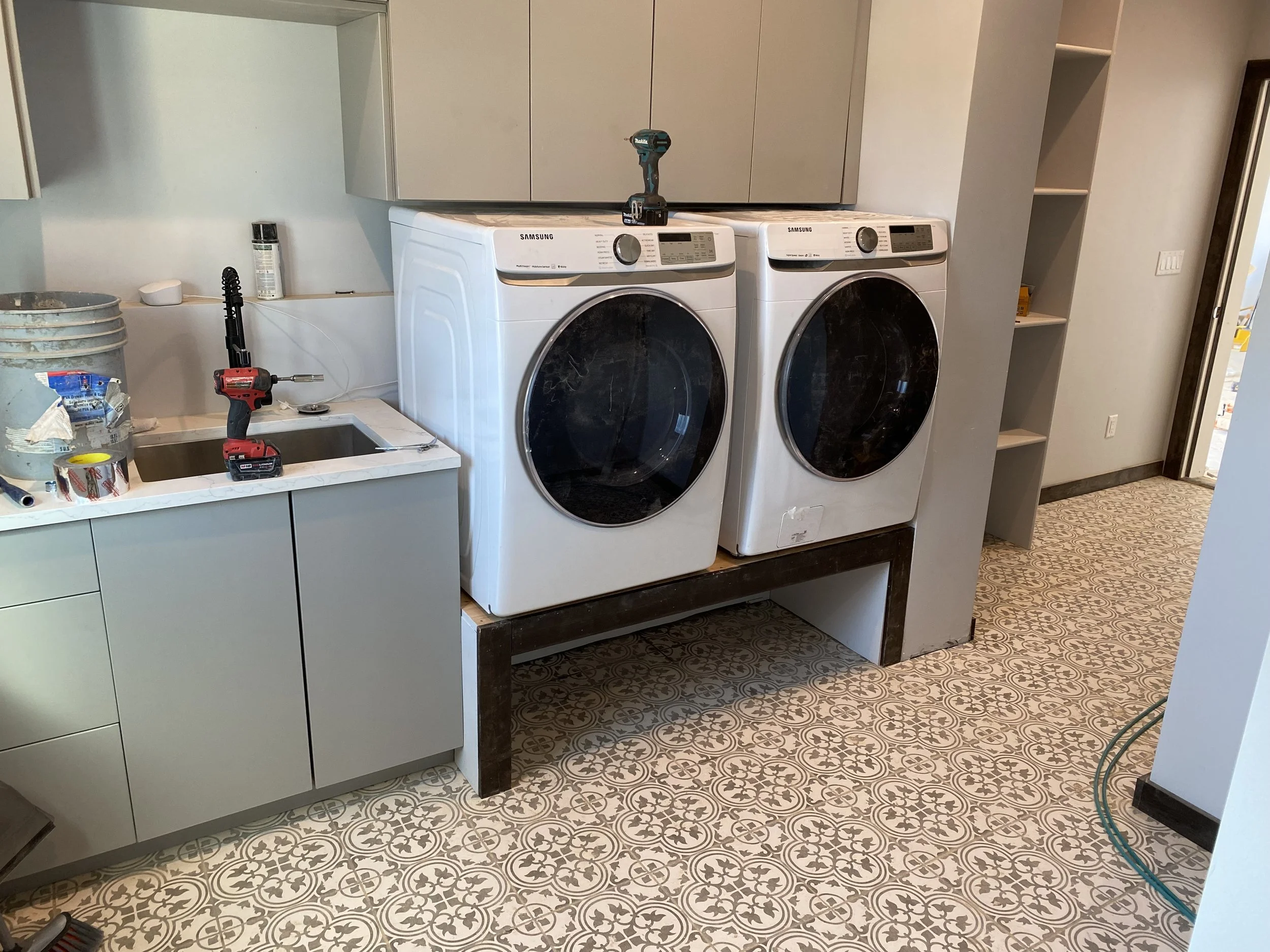 Laundry room with a washing machine and dryer on a raised platform, a countertop with a sink, tools including a cordless drill, and construction materials.