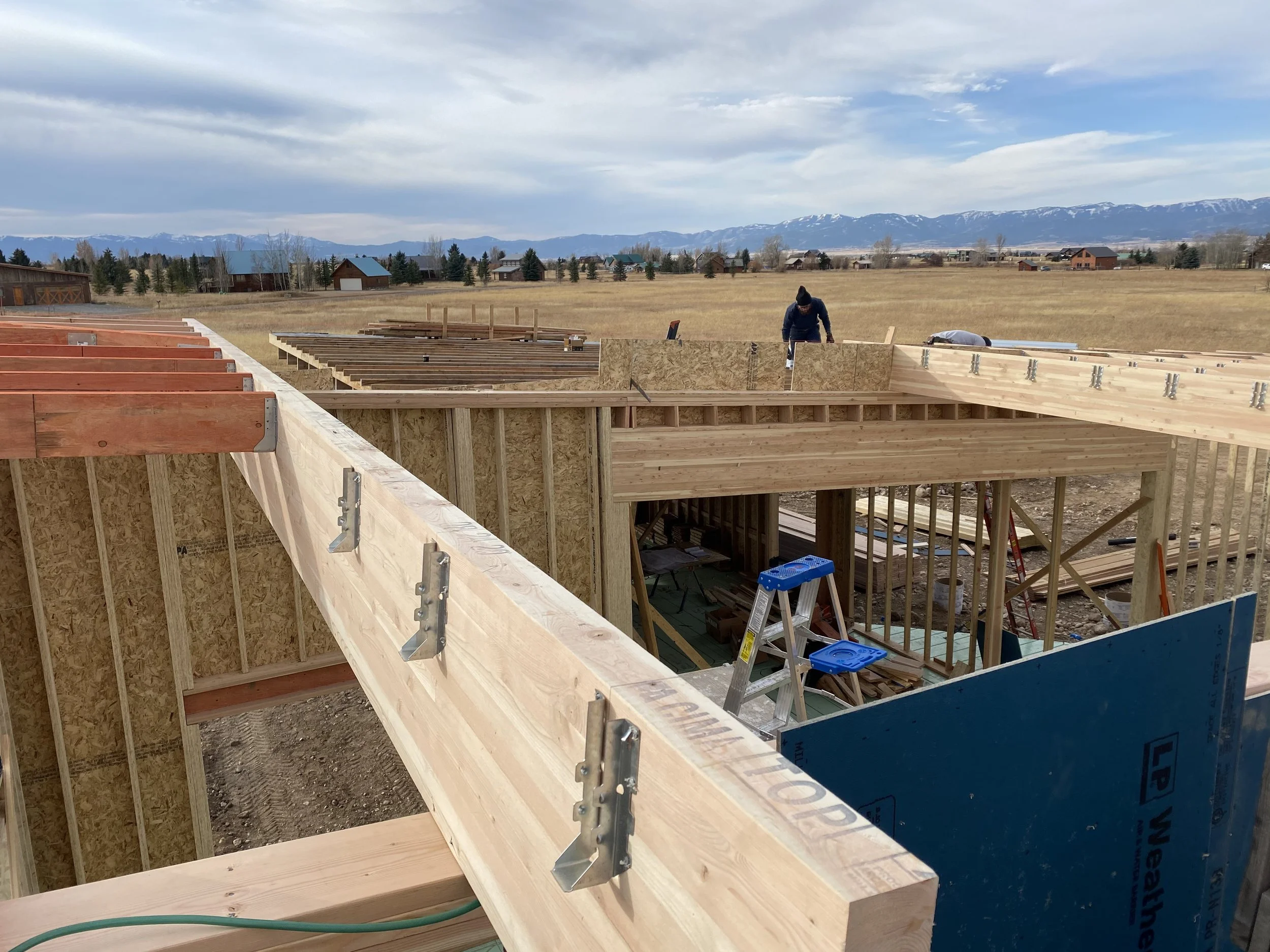Construction site with wooden framing and workers building a house in a rural area with mountains in the background.
