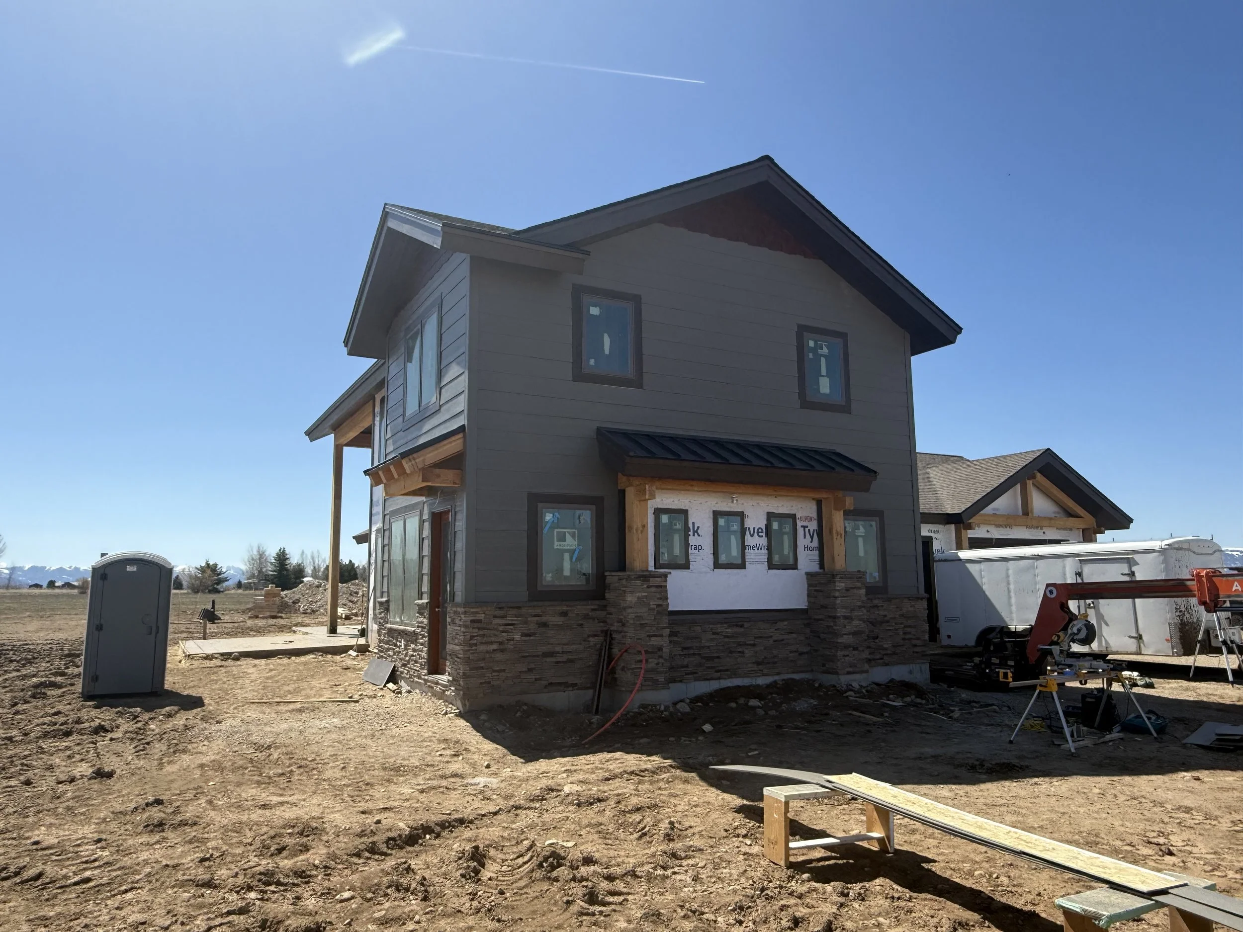 A house under construction in a rural area with clear blue sky, construction equipment, and building materials around.