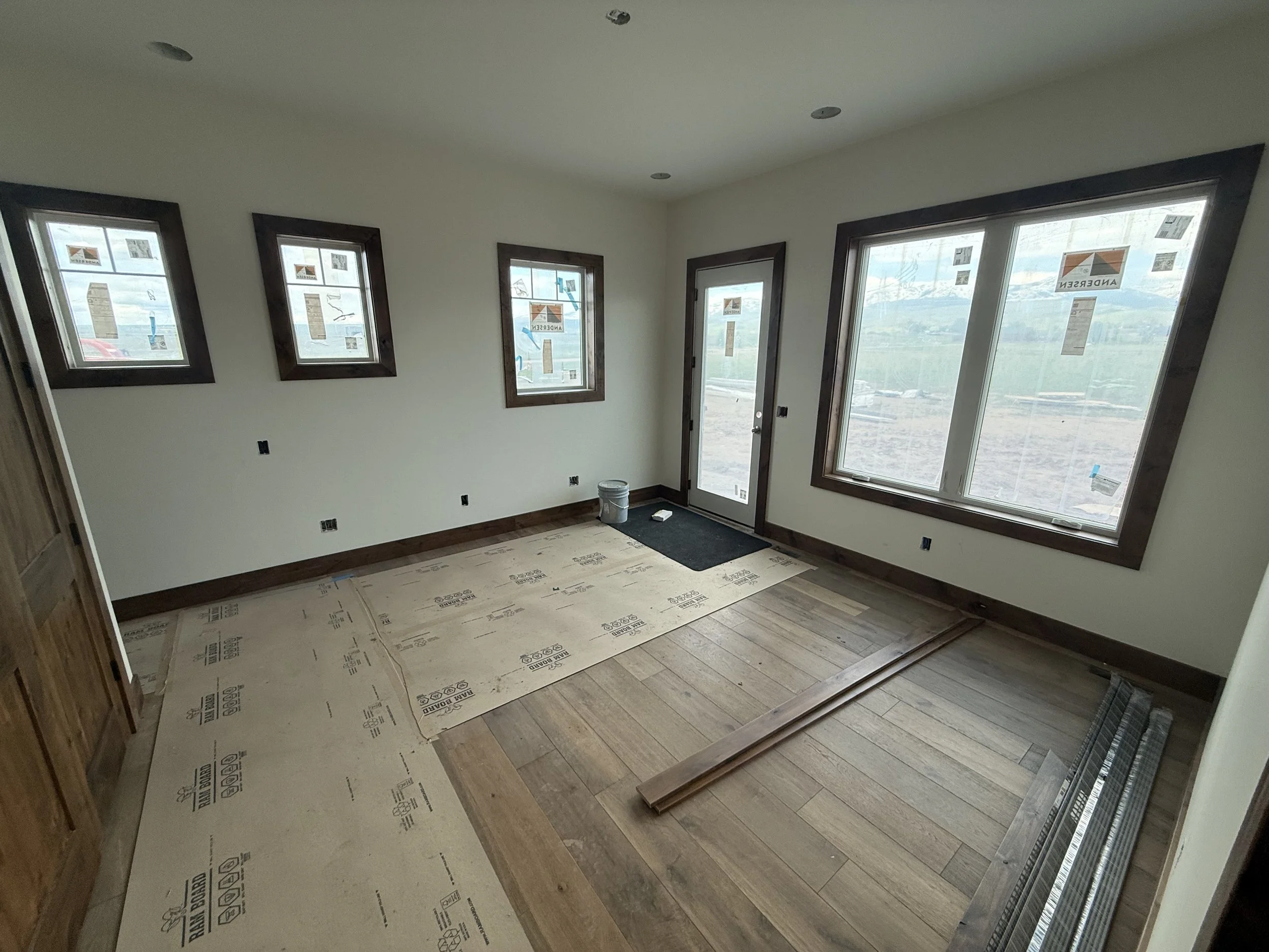 Interior of a room under construction with new wooden flooring, four windows, a glass door, and construction materials on the floor.