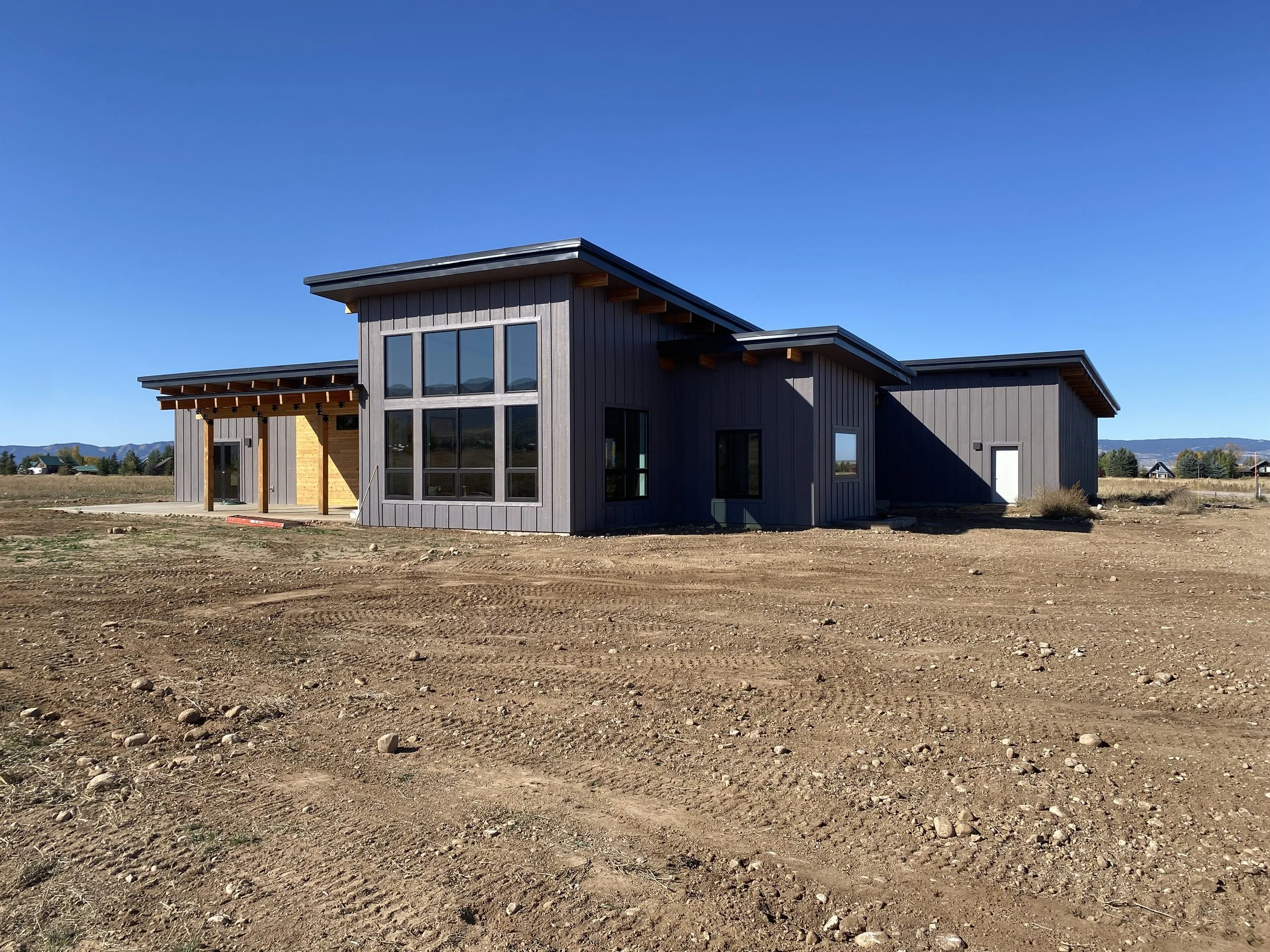 Construction site with a modern, gray house featuring large windows and a covered porch, in a rural area with dirt ground and a clear blue sky.