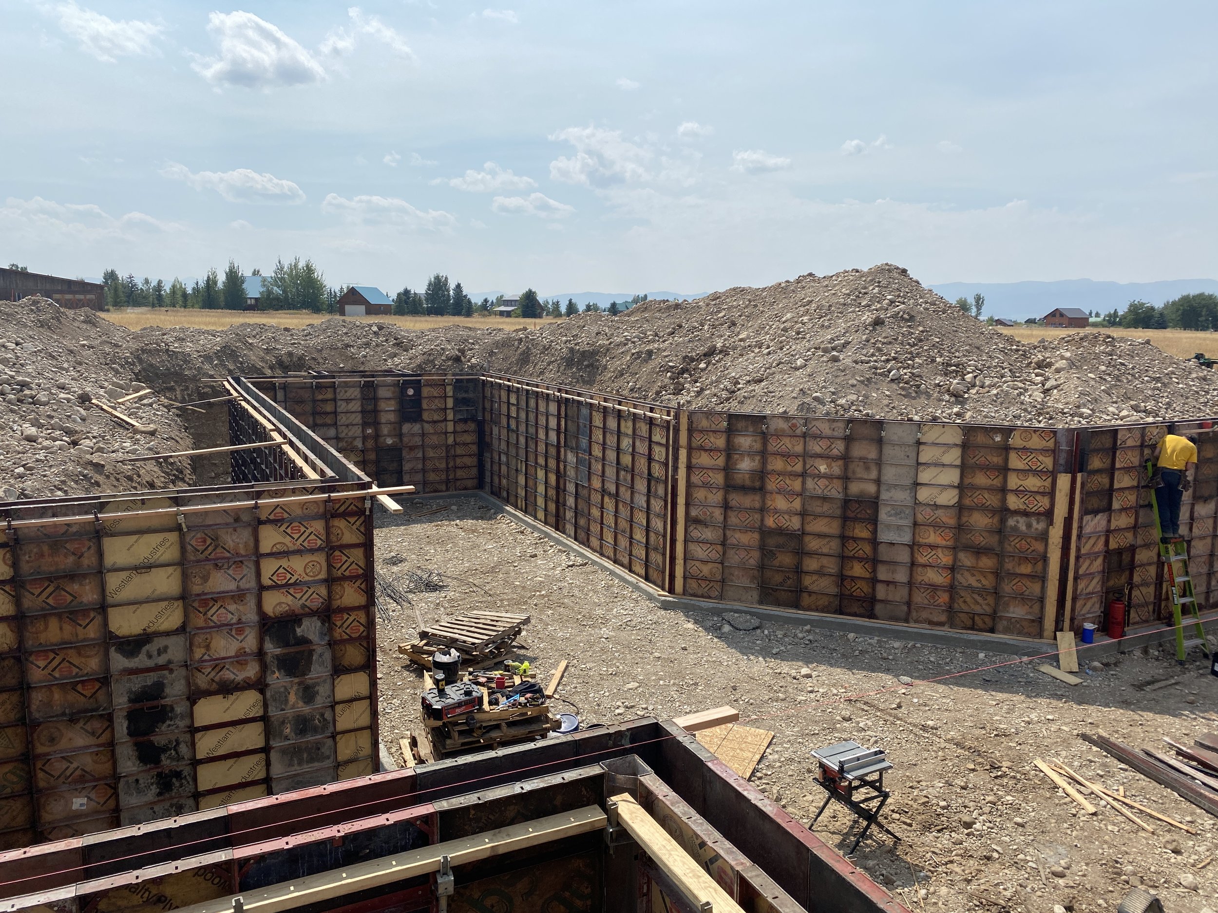 Construction site with concrete formwork walls, a worker on a ladder, and piles of dirt in a rural area with fields and houses in the background.