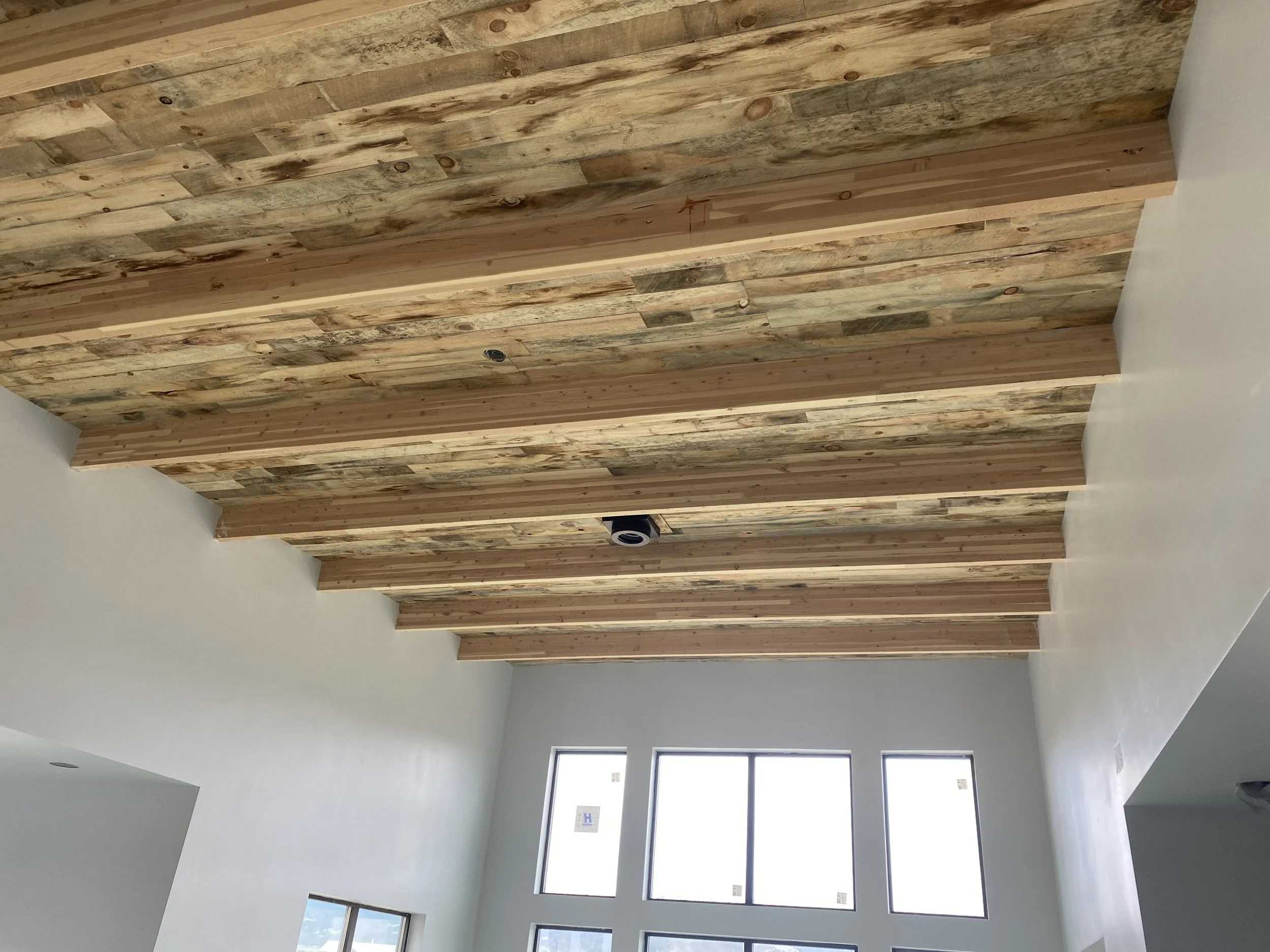 Interior room with a wooden ceiling made of planks and beams, white walls, and large windows letting in natural light.