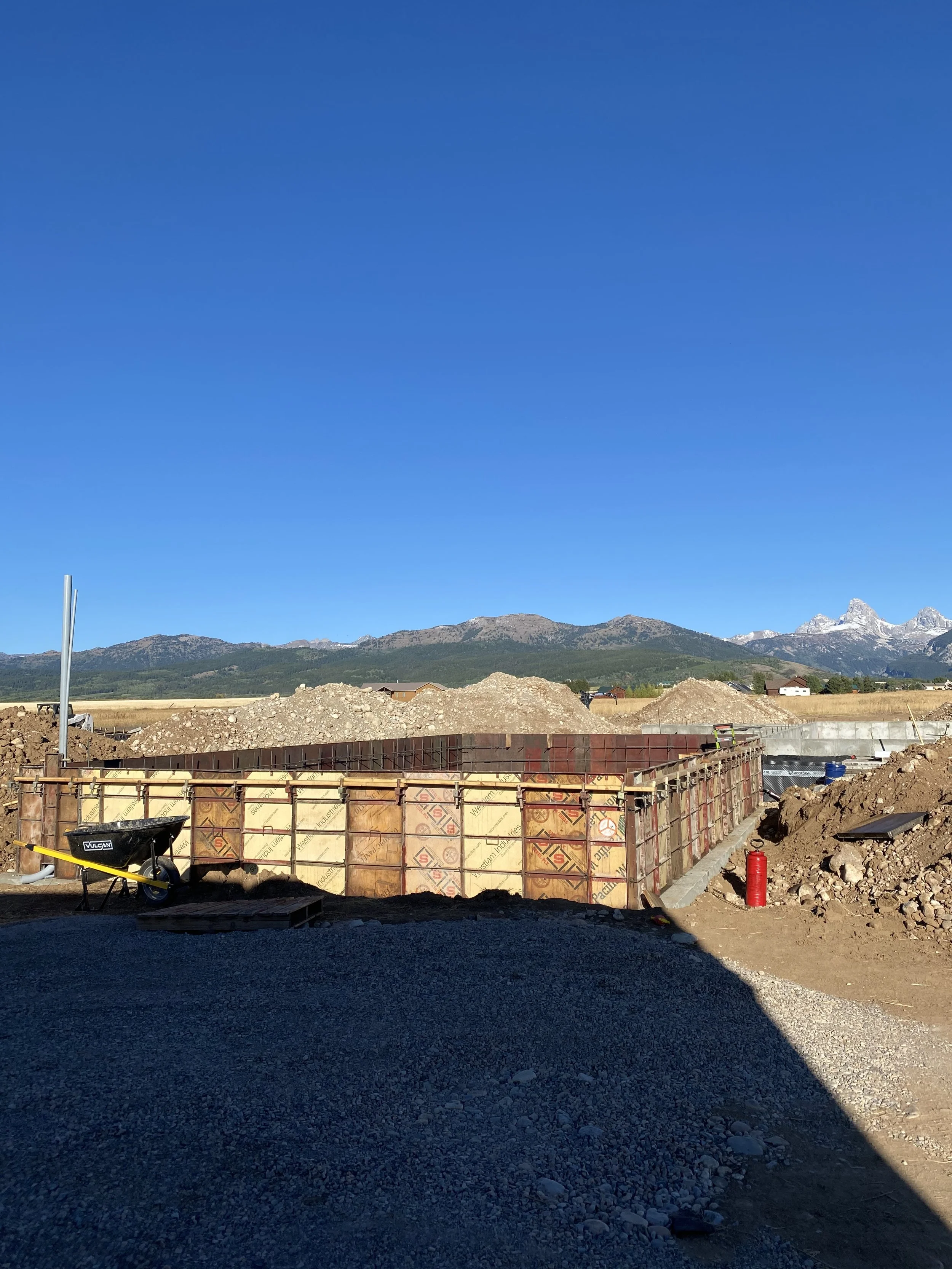 Construction site with concrete formwork, a wheelbarrow, and a fire extinguisher, with mountains in the background and a clear blue sky.