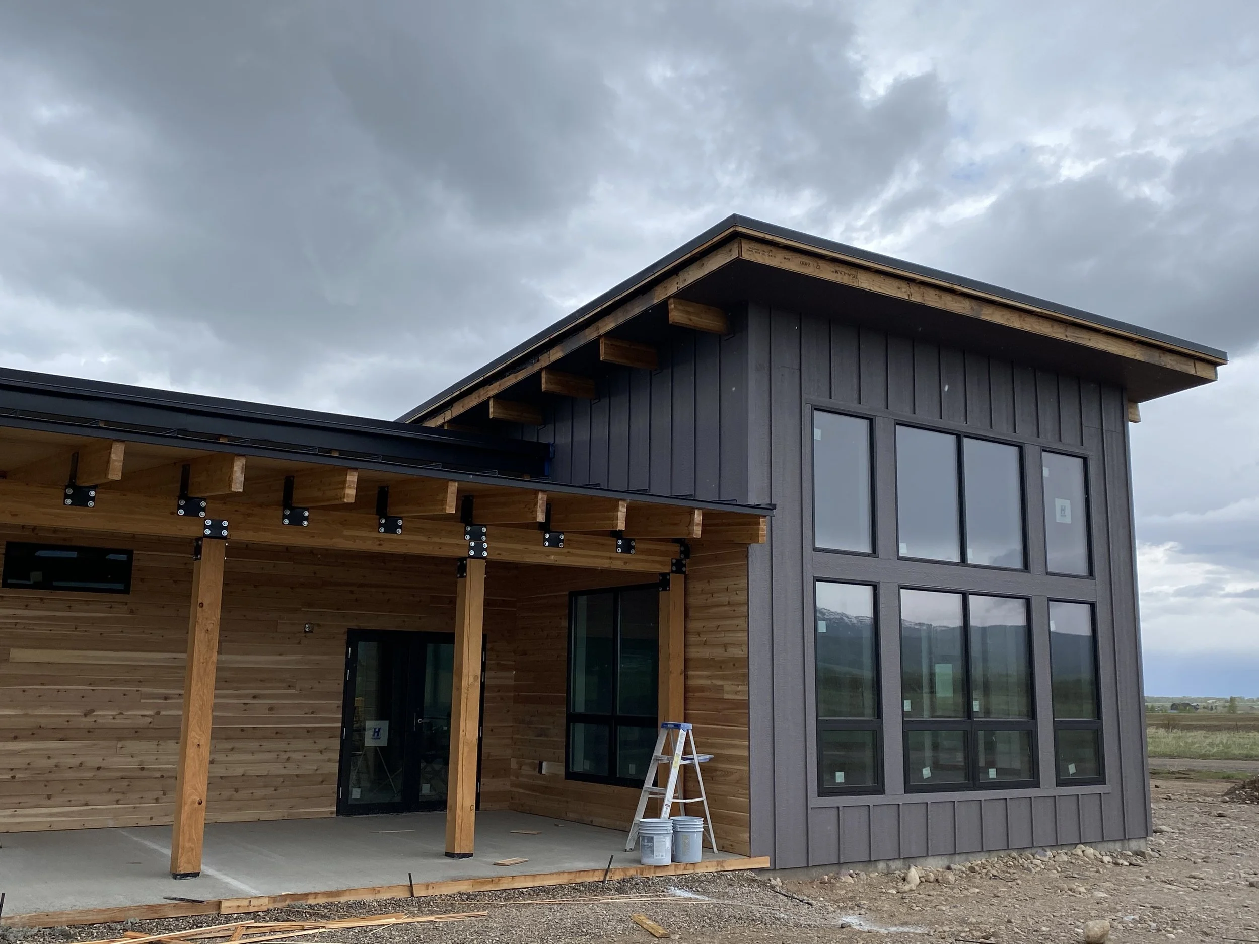 Custom home entry with timber framing, large windows, and natural wood accents in Teton County, Idaho.
A modern two-story house under construction with mixed wood and gray exterior, large windows, and an overhang roof under cloudy skies in Driggs Ida