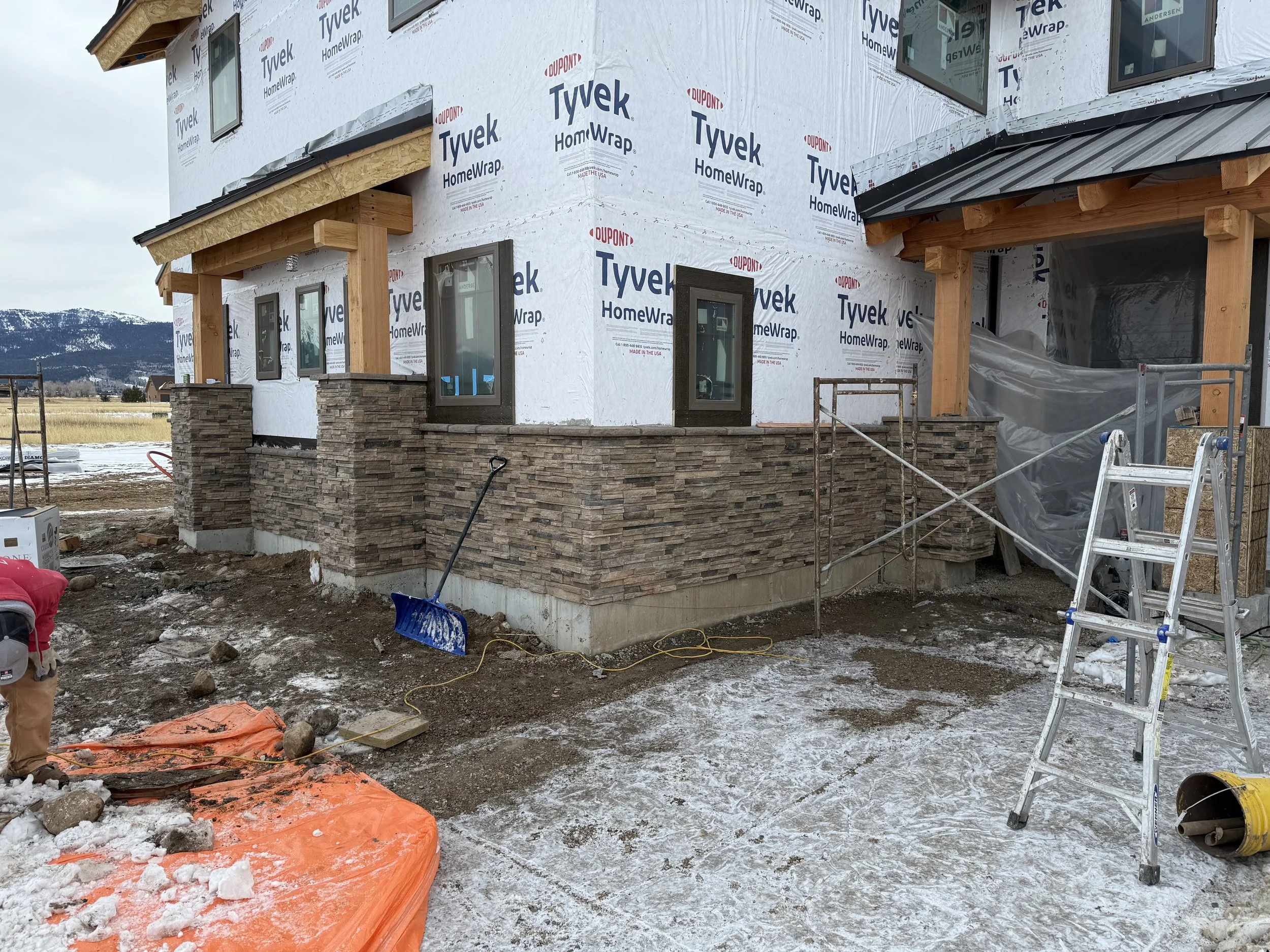Exterior of a house under construction with brick and stone siding, several windows, and wooden beams. Construction materials and tools are visible on the snowy ground.