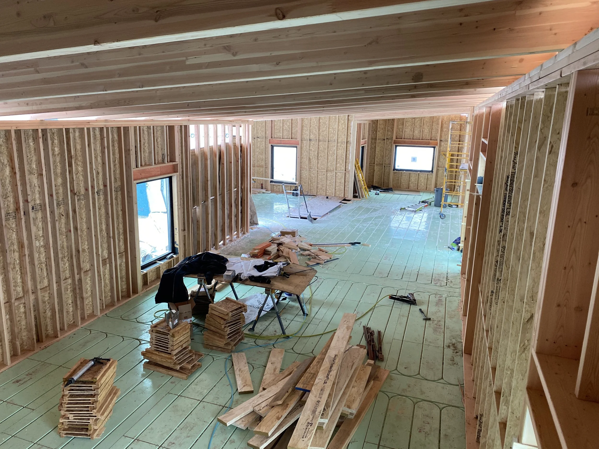 Interior of a house under construction with exposed wooden studs, green insulation on the floor, and construction tools and materials scattered around.