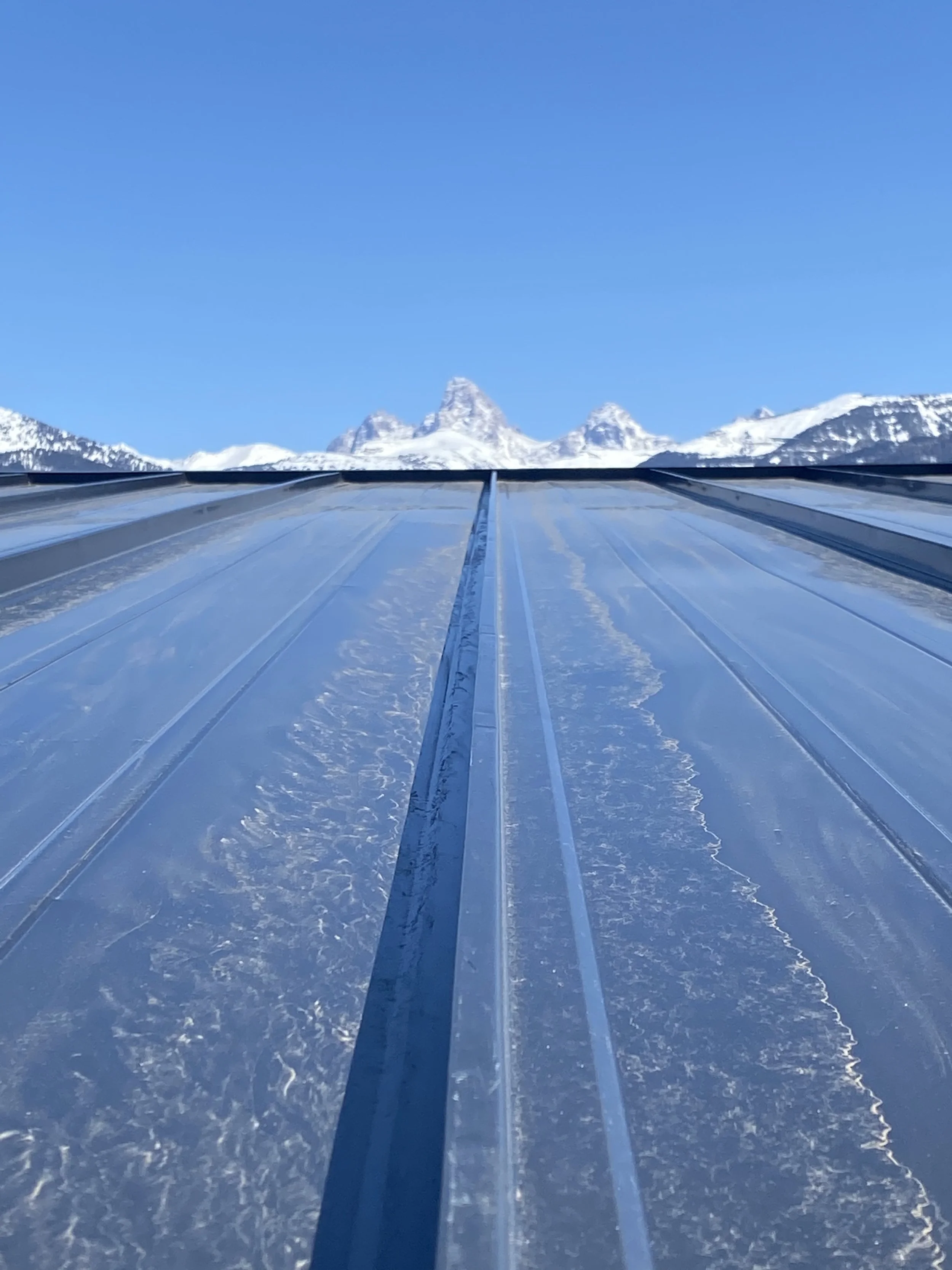 Snow-covered metal roof with mountain peaks in the background under a clear blue sky.