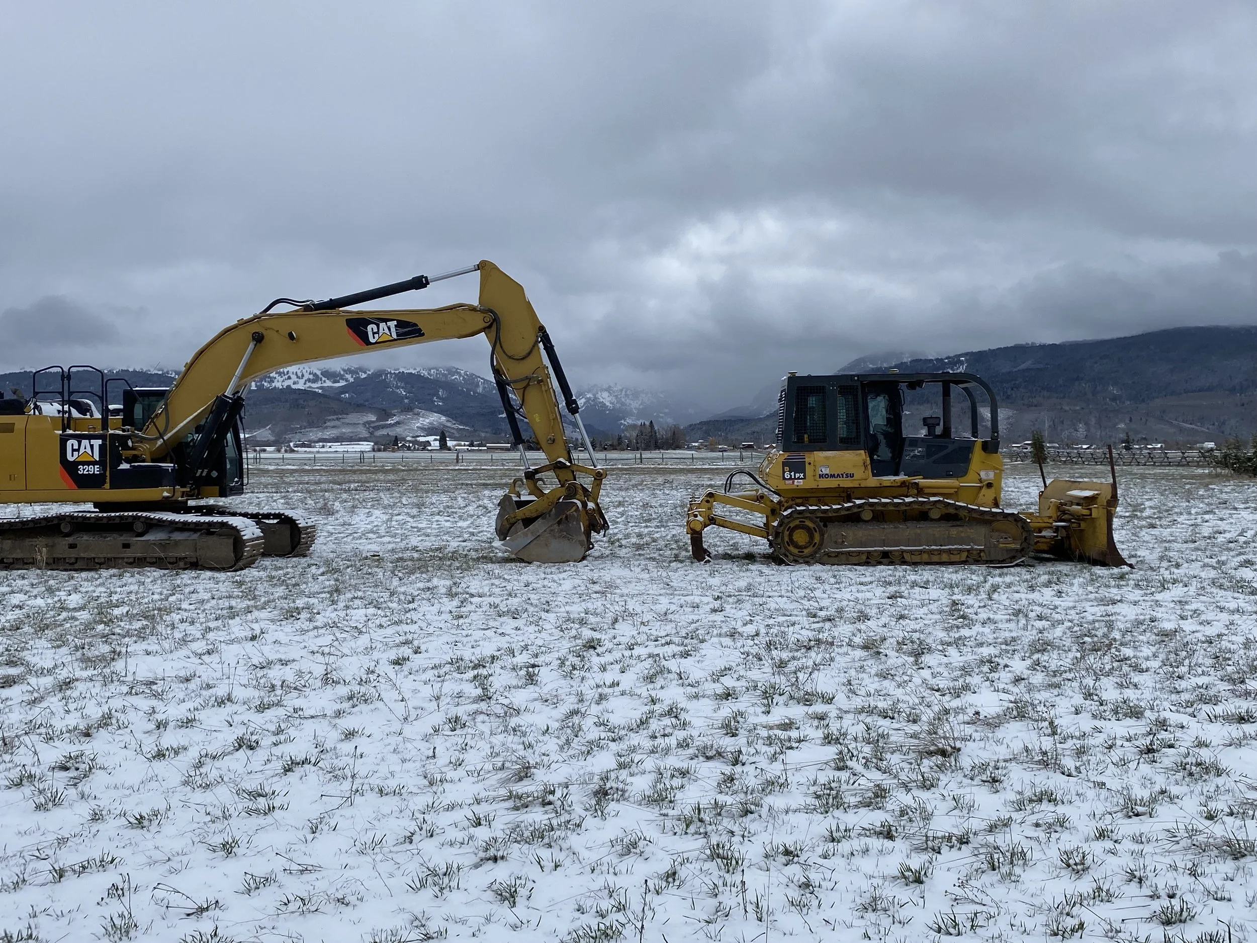 Two yellow construction vehicles, a large excavator on the left and a smaller bulldozer on the right, parked on a snow-covered field with mountains in the background and a cloudy sky overhead.