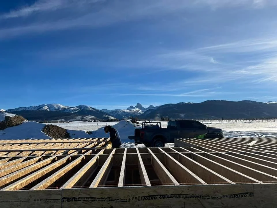A person working on the framework of a building under construction in a snowy landscape with mountains in the background.