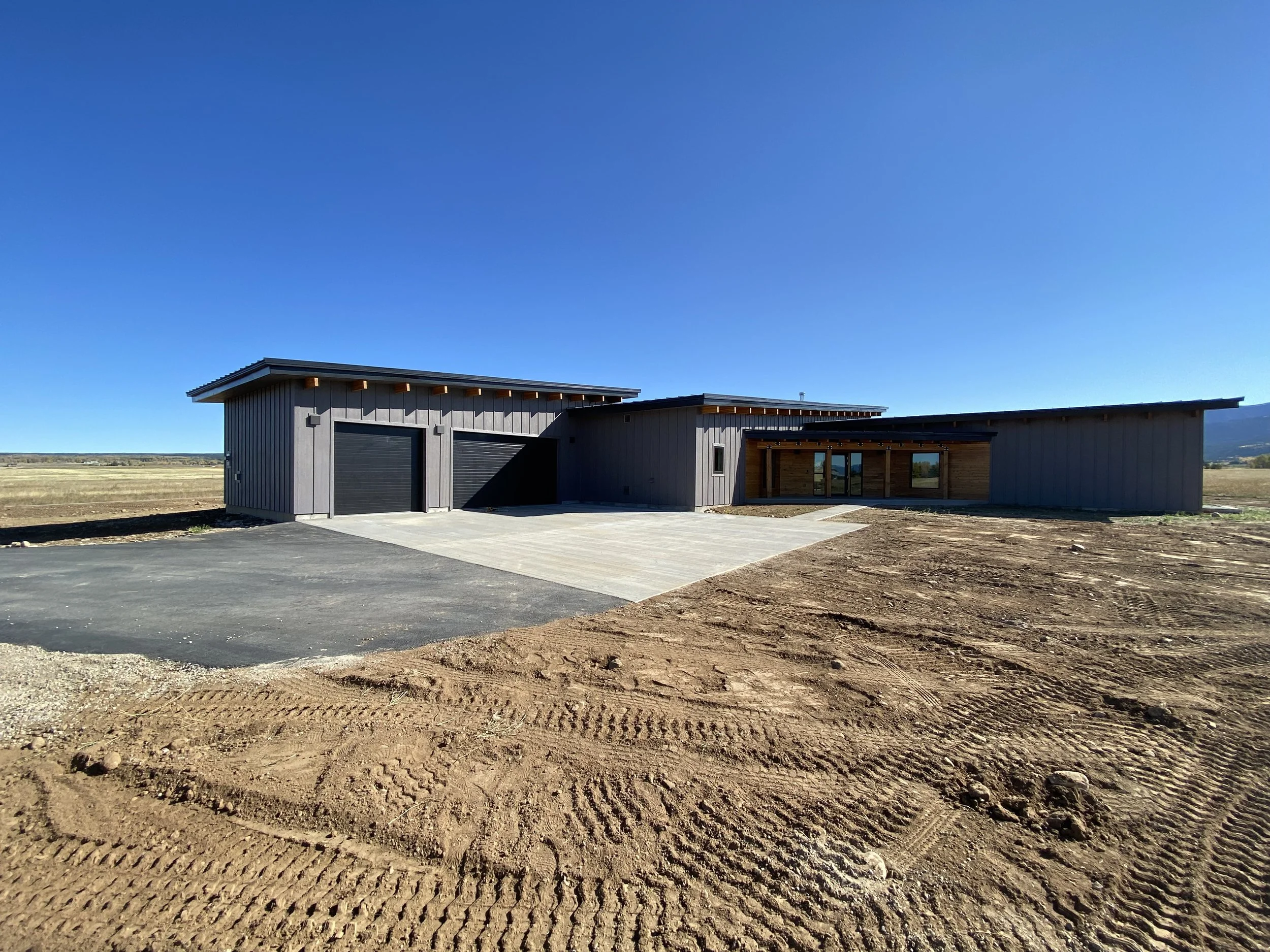 Custom home with large driveway and modern exterior design built in Eastern Idaho. Modern house with black garage doors and a wooden overhang, situated on a construction site with dirt and tire tracks in the foreground and a clear blue sky.