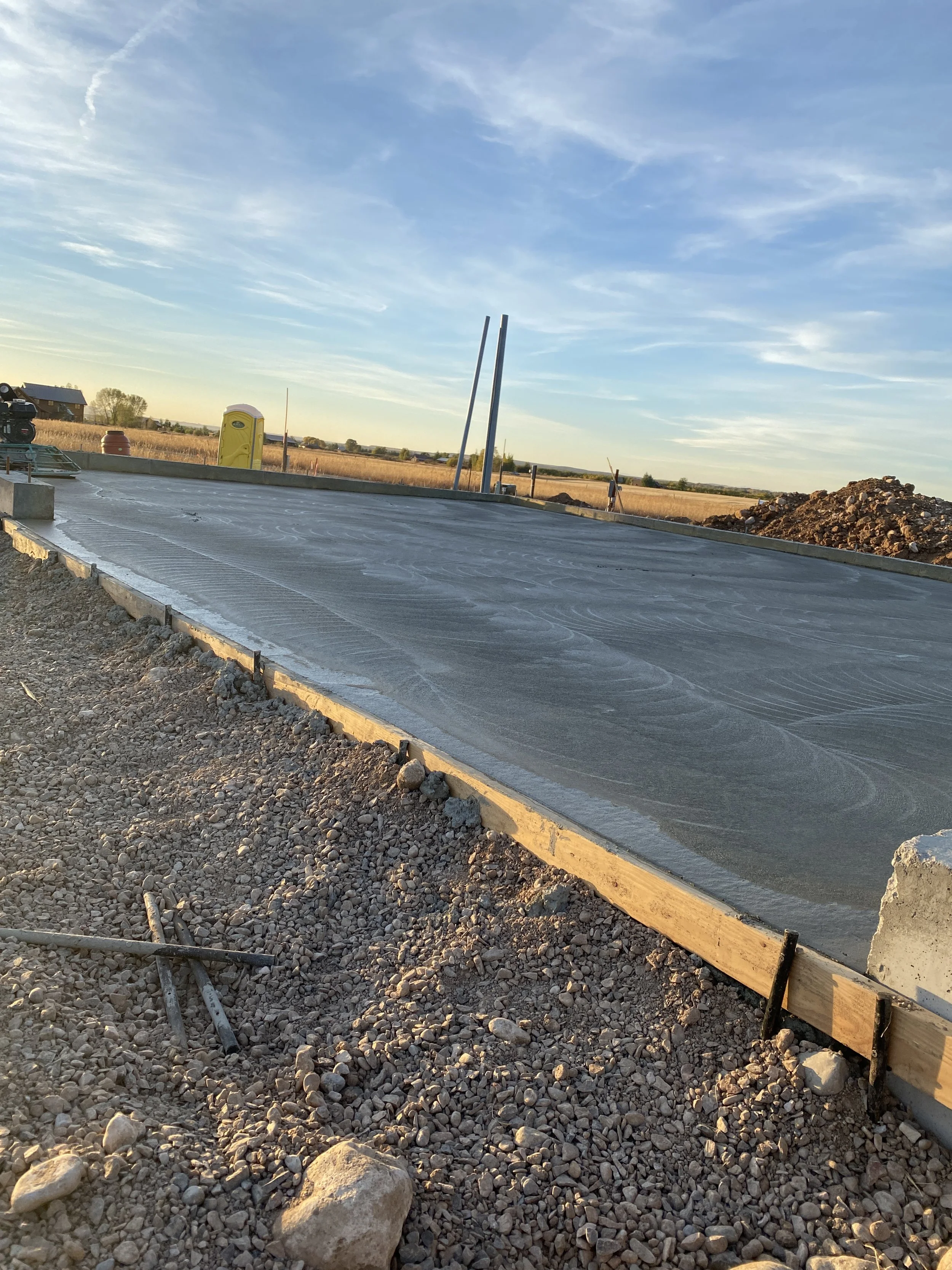 A freshly poured concrete slab on a construction site, surrounded by gravel and wooden forms, with a yellow portable toilet and a blue sky with wispy clouds in the background.