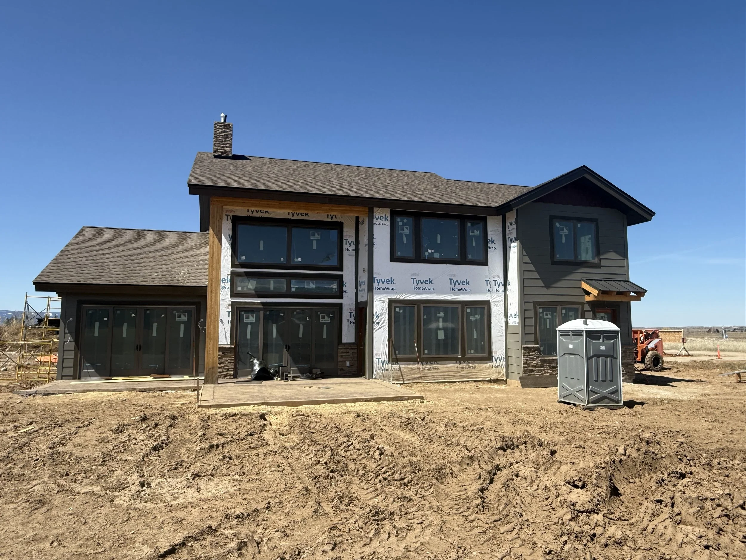 A two-story house under construction, with partially finished exterior walls, large windows, and a small porch area, set on a dirt lot on a clear day.