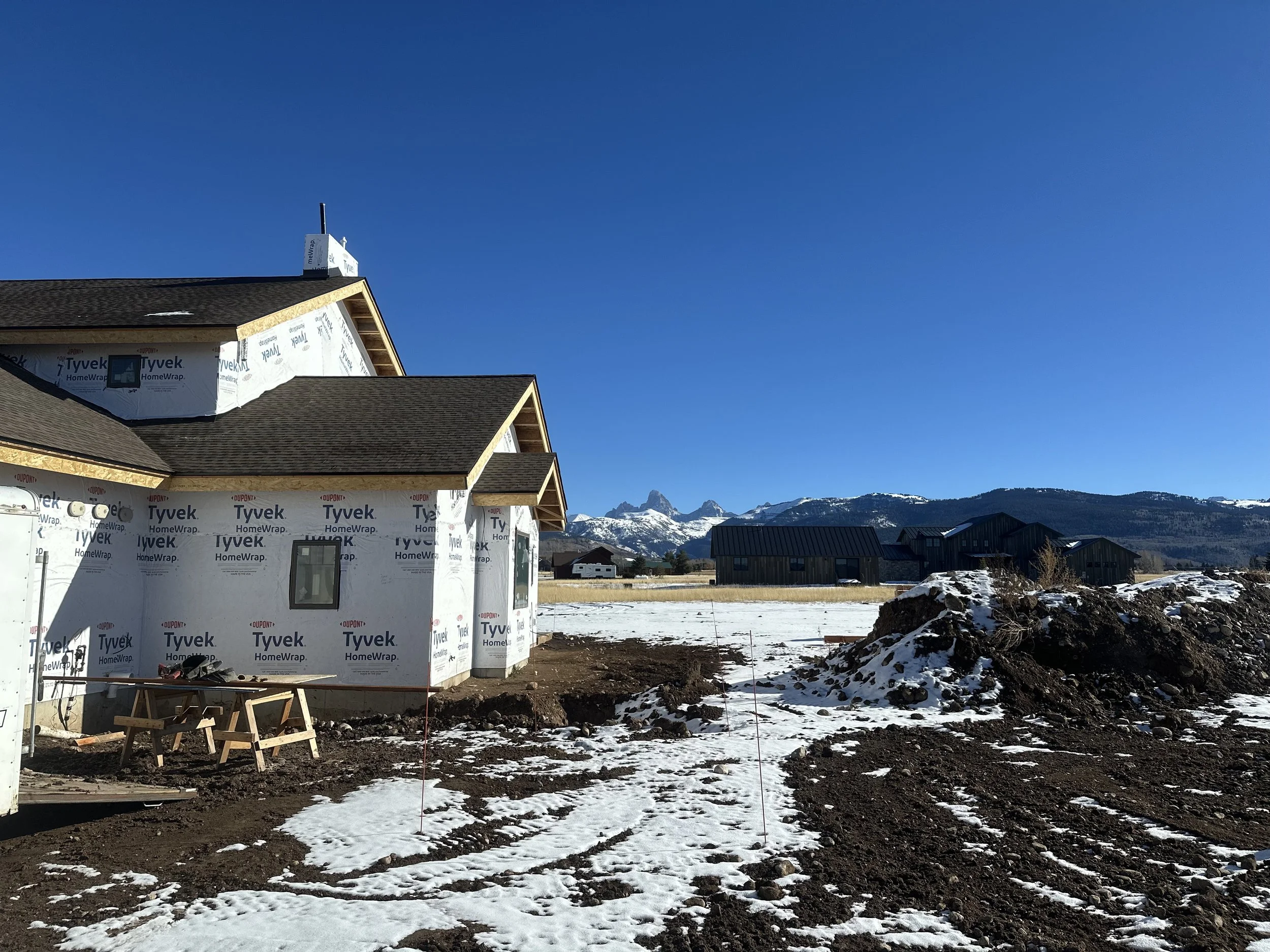 A house under construction with Tyvek HomeWrap exterior sheathing, snow on the ground, and a mountain range in the distance under a clear blue sky.