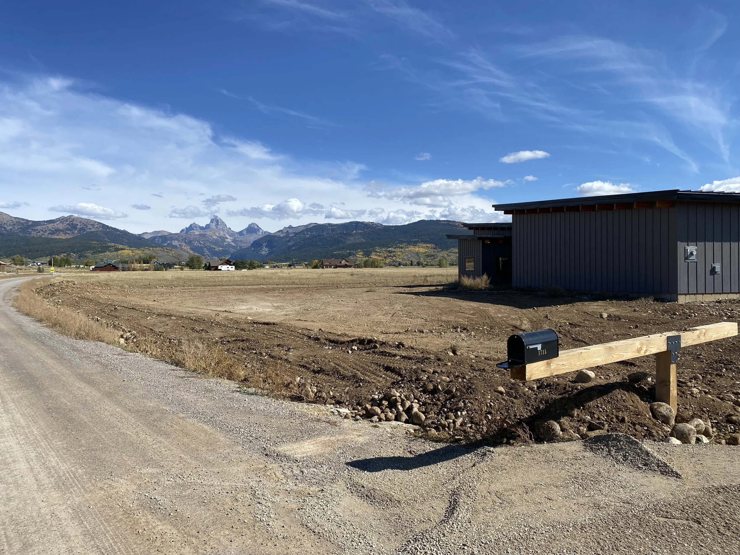 A rural scene with a dirt road on the left, an open field, a modern gray building on the right, a black mailbox on a wooden post in the foreground, and mountains with rocky peaks in the background under a partly cloudy blue sky.