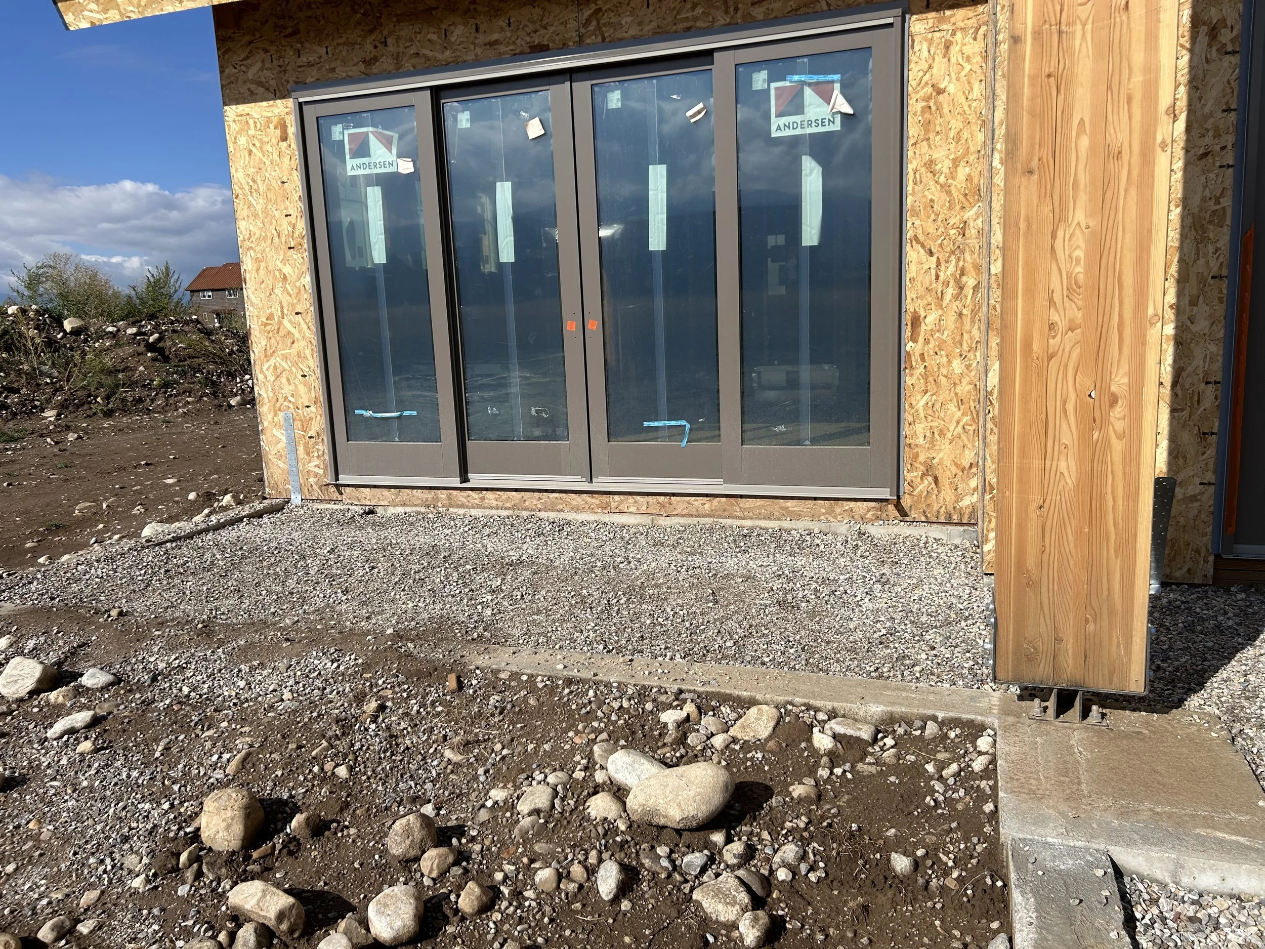 Exterior view of a building under construction with a large sliding glass door framed in gray, set in a wooden structure with plywood sheathing. The ground in front is gravel and dirt with scattered rocks.