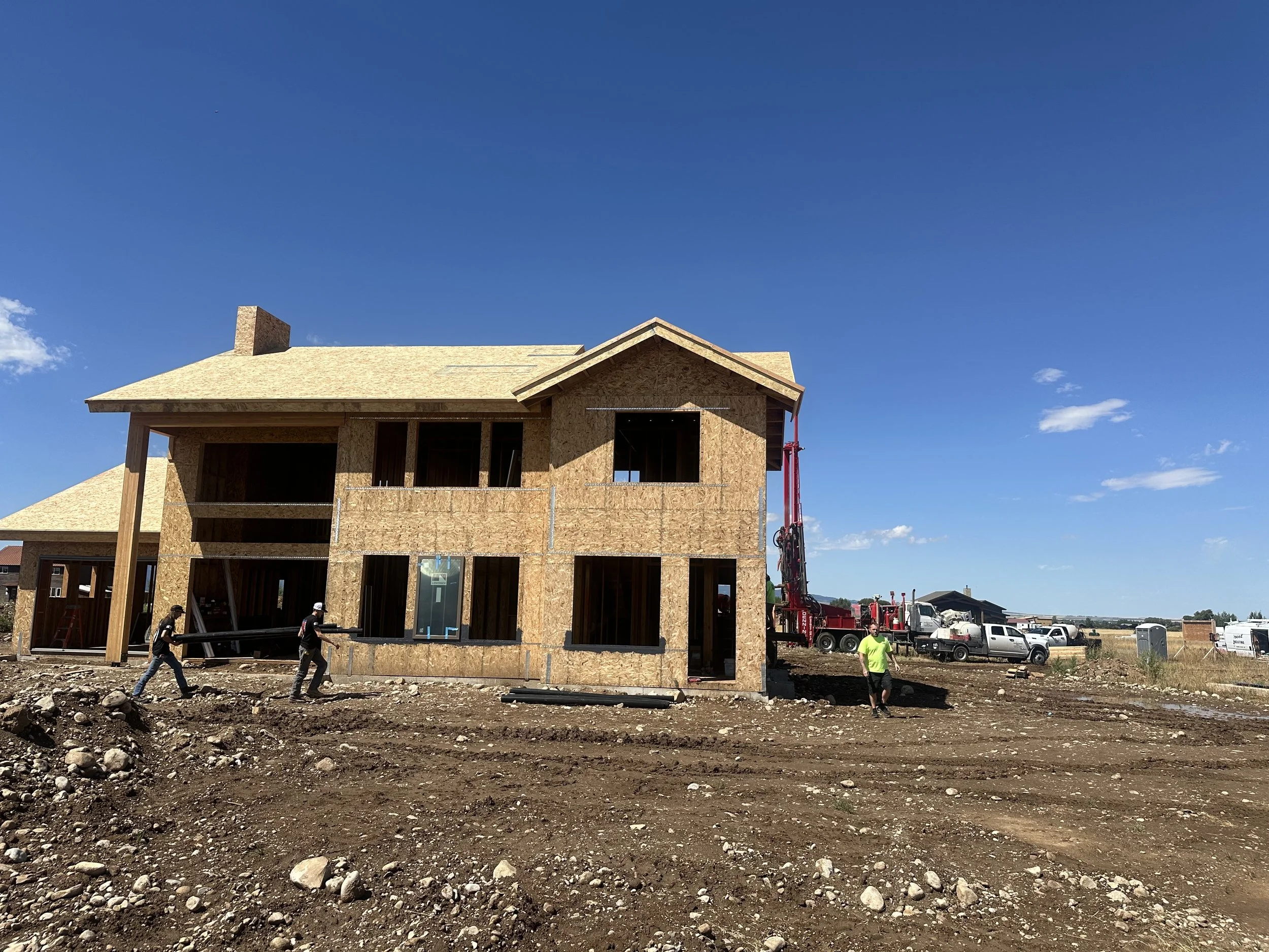 Construction site with a partially built two-story wooden house, workers, and construction equipment under a blue sky.