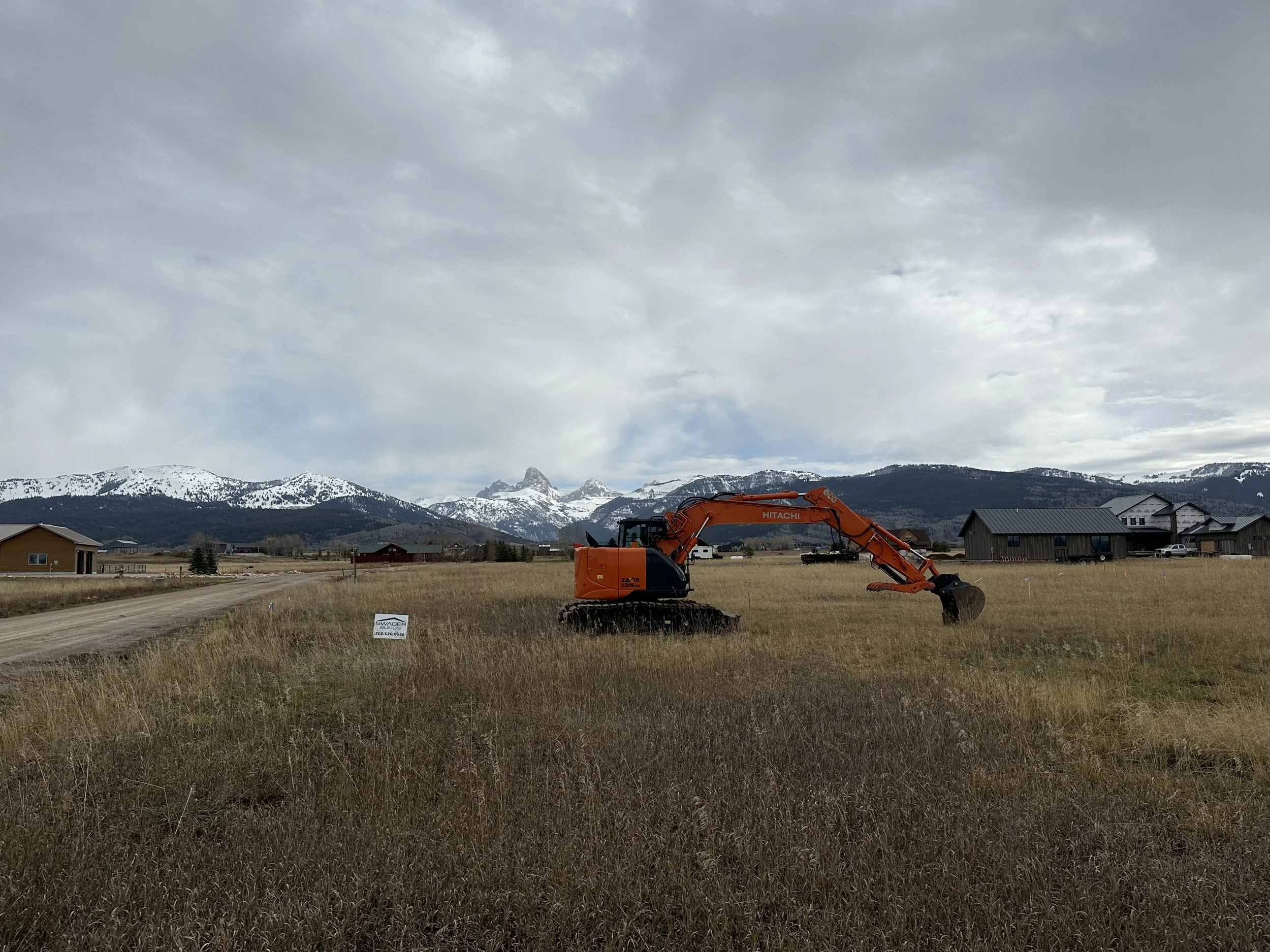 A bright orange Hitachi excavator operating in a field with houses and snow-capped mountains in the background under a cloudy sky.