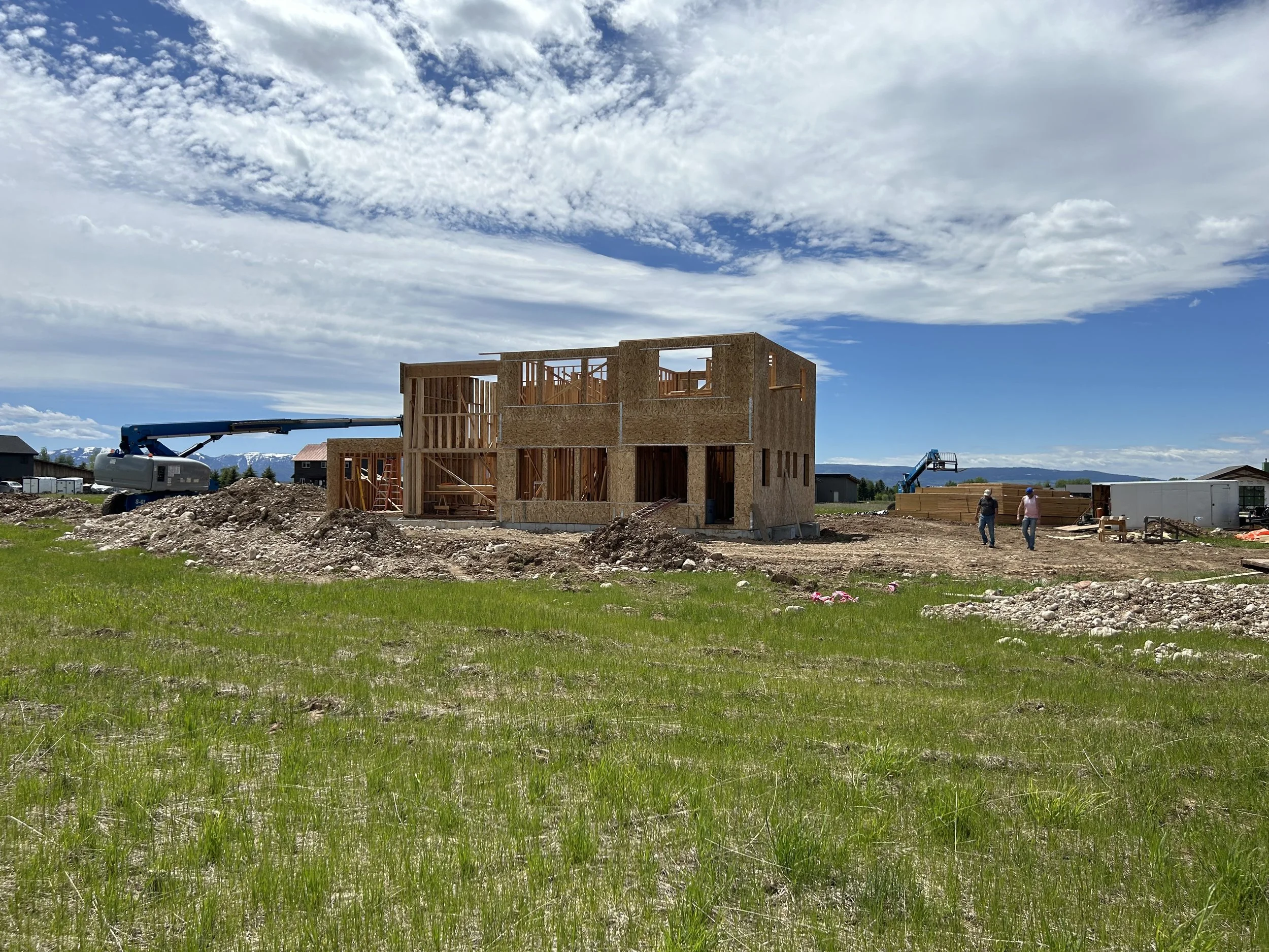 A wooden house under construction on a grassy field, with construction equipment and workers nearby, under a partly cloudy sky.