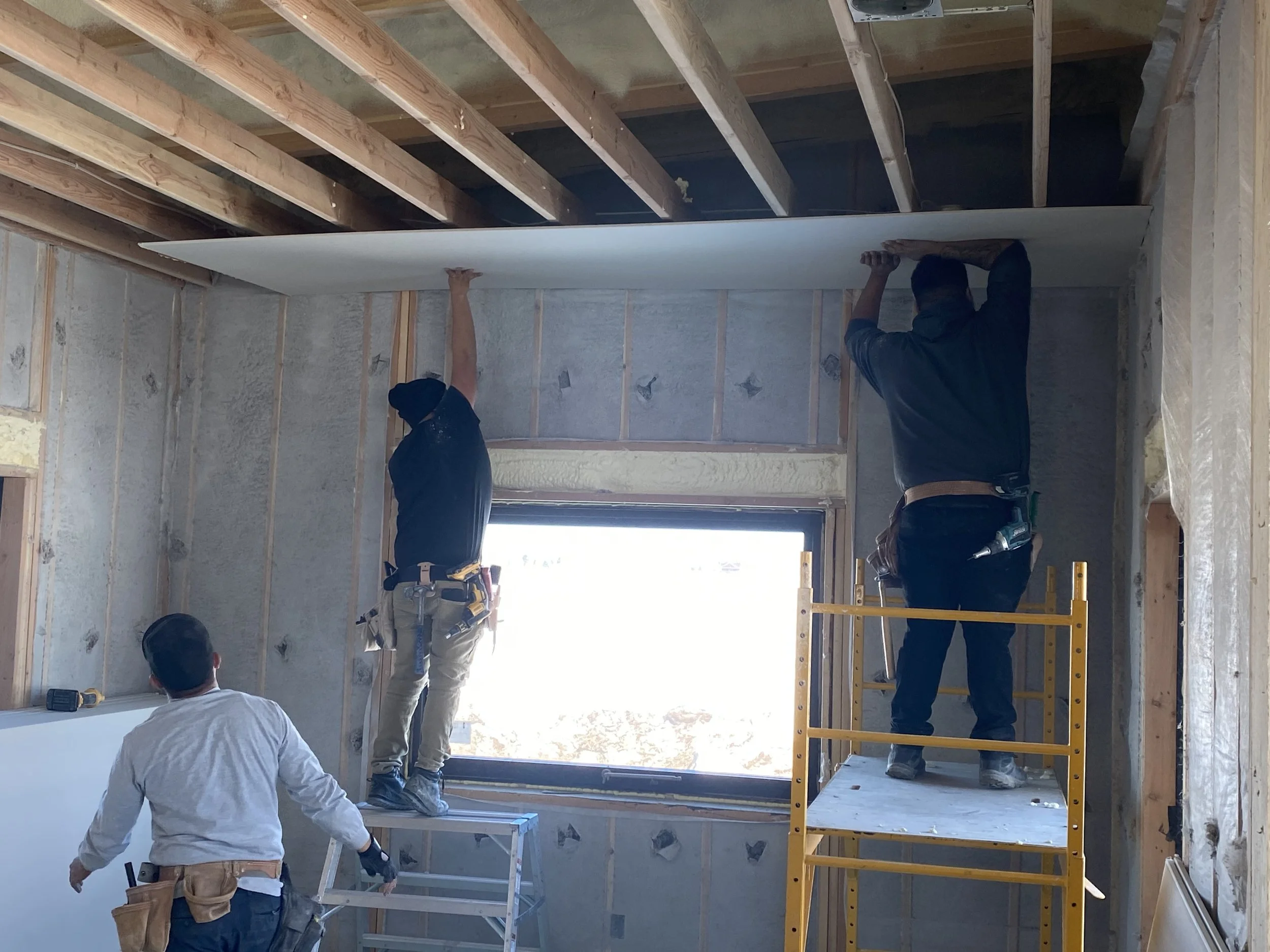 Three construction workers installing drywall inside a building under renovation. Two workers are standing on scaffolding, attaching a large sheet of drywall to the ceiling, while the third worker stands on a step ladder, working on the wall.