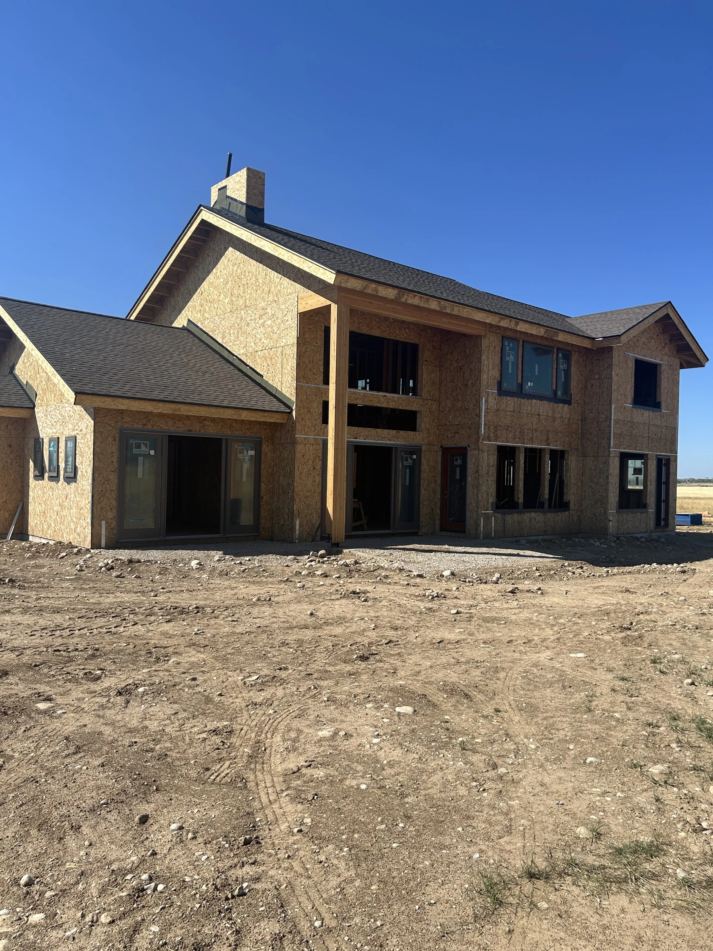 Under construction two-story house with plywood exterior, multiple windows, and a pitched roof, on a dirt lot under a clear blue sky.