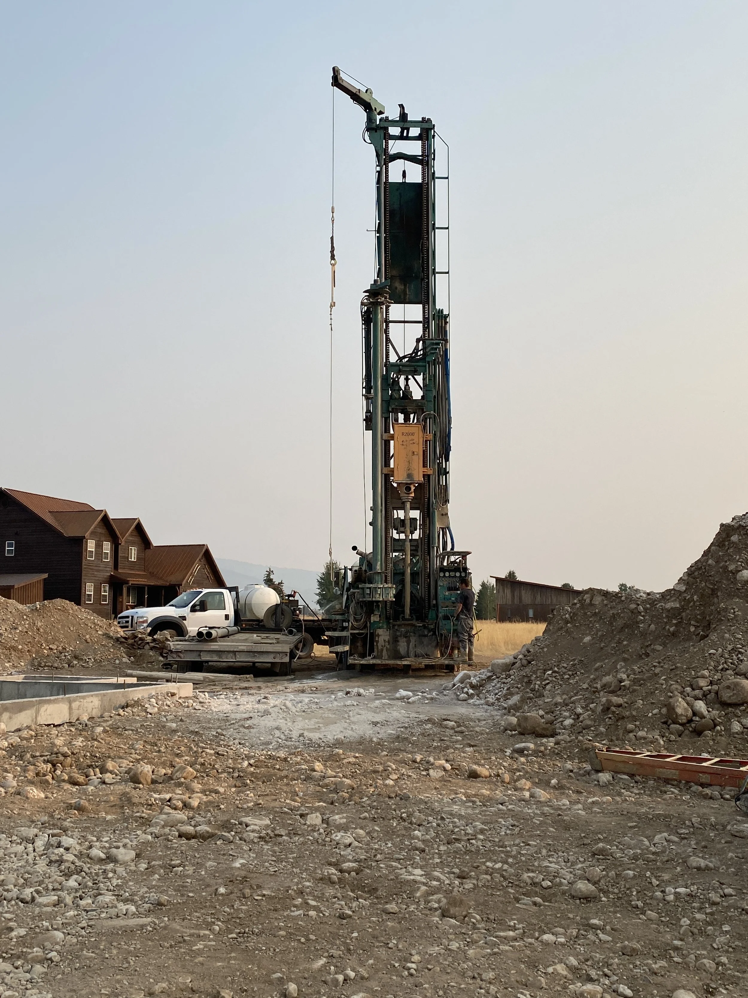 Construction site with tall drilling machine, pickup truck, and piles of dirt and rocks.