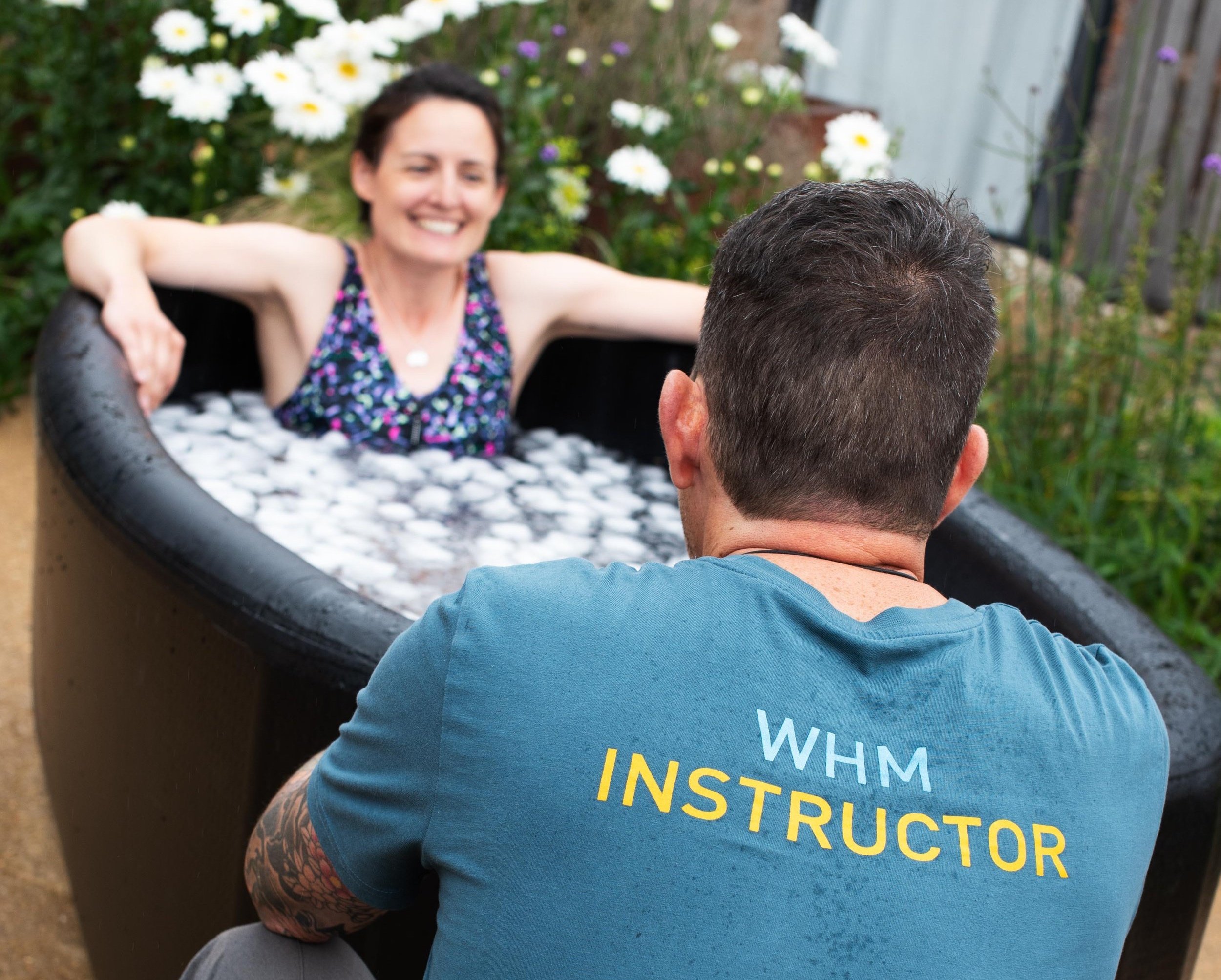 A smiling woman in a colorful sleeveless top sitting in a hot tub, with a man sitting in front of her, facing away, outdoors with flowers and a fence in the background.