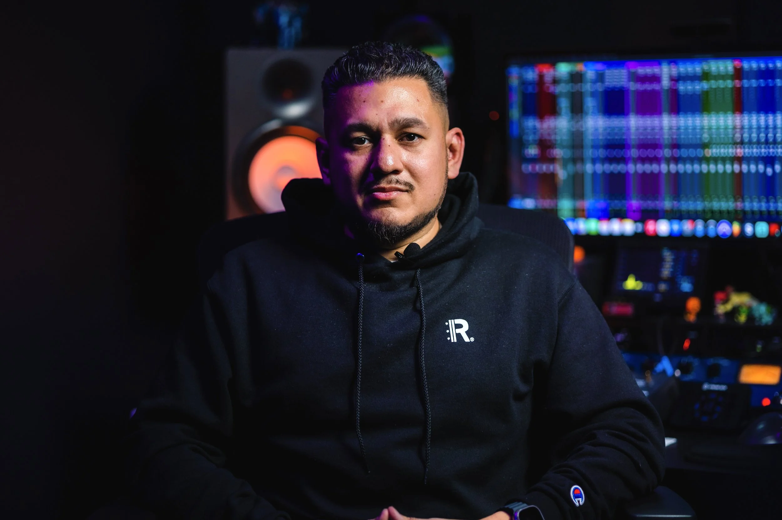 A man with short dark hair and a beard sitting in a dimly lit room in front of a computer monitor with colorful audio editing software, wearing a black hoodie.