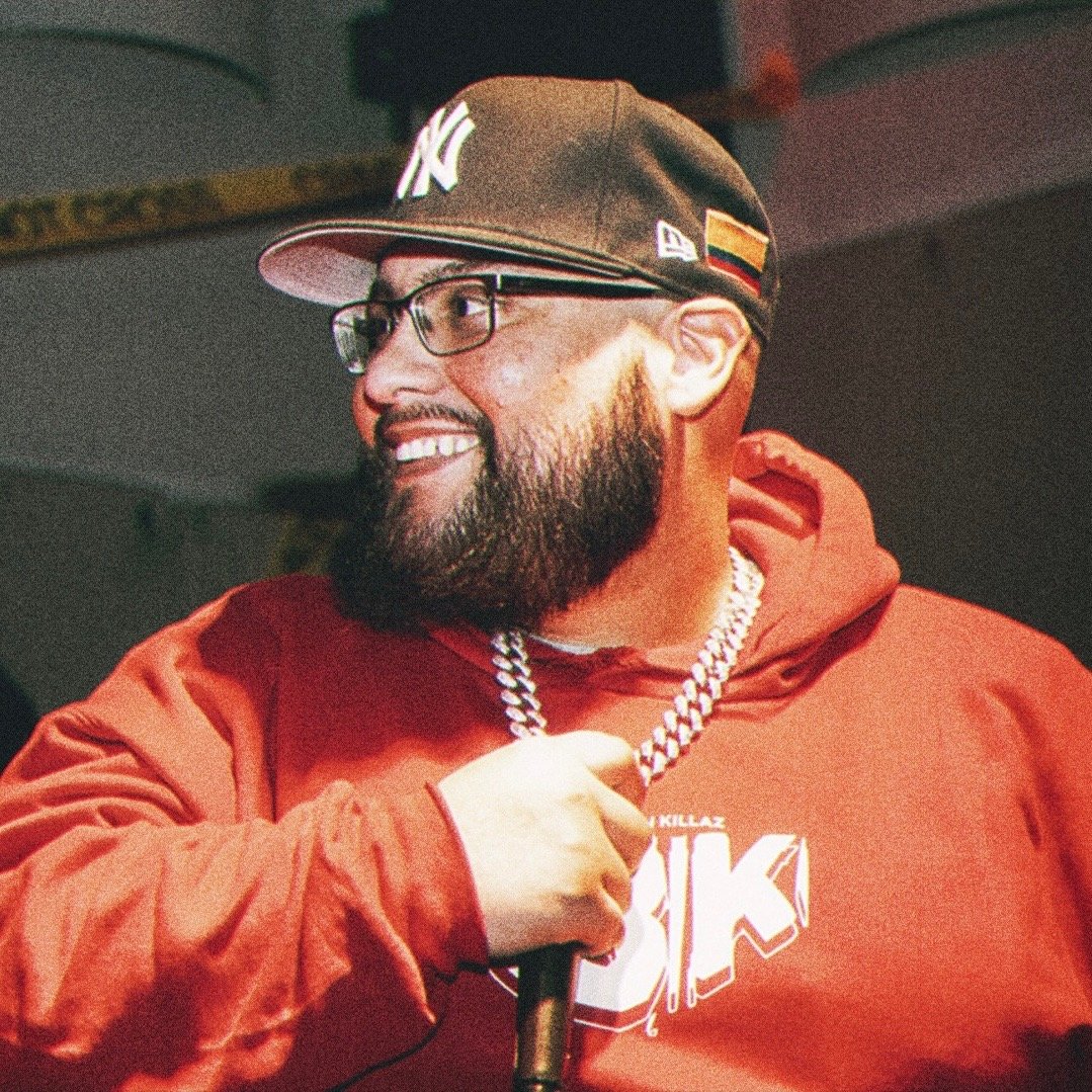A man with a beard, glasses, and a baseball cap smiling while holding a microphone. He is wearing a red hoodie and a diamond chain, with a flag on his cap and a logo on his hoodie.